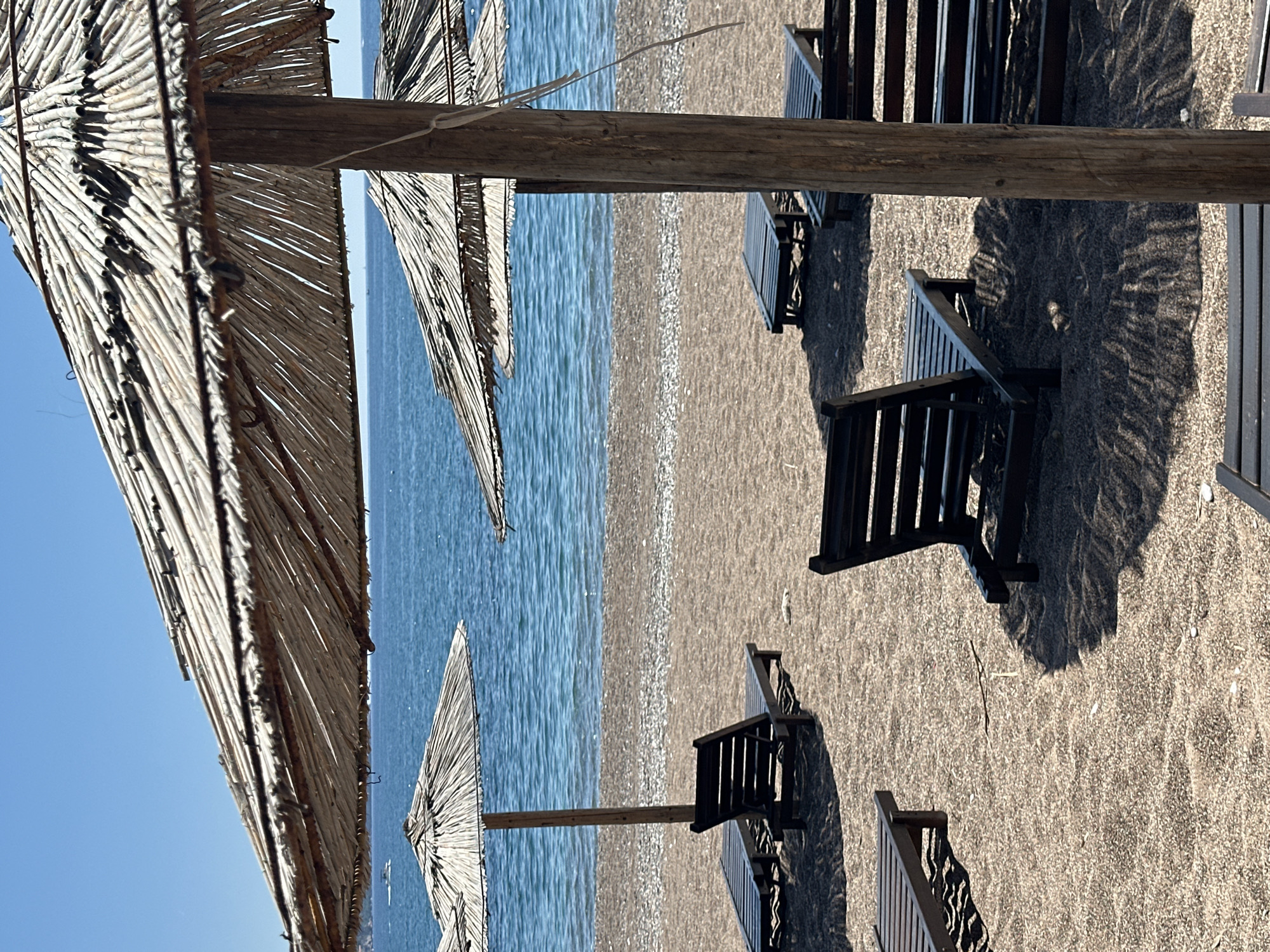 The image shows a beach scene in Boreti, Montenegro. In the foreground, there are several thatched umbrellas providing shade to empty dark-colored beach chairs. The beach consists of light-colored sand and pebbles. Beyond the beach is a calm, blue body of water, and in the background, a hazy sky is visible. The scene lacks any signs of human activity.