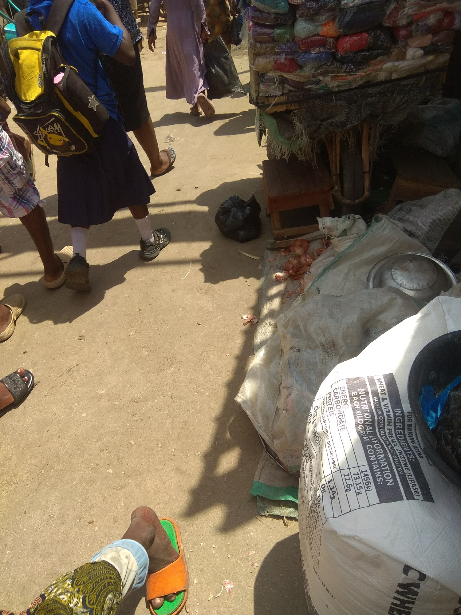 The image depicts a busy outdoor market scene in Jalingo, Nigeria. In the foreground, a child wearing a blue shirt, a dark blue skirt, white socks, and black sneakers is walking to the left, carrying a yellow and black backpack adorned with a Pikachu character. To the child's left, another person's legs are visible, wearing checkered shorts and sandals. Further to the left, the legs of a person wearing a patterned fabric and blue pants, and an orange sandal are visible.
In the background, a person in a light purple dress walks away from the camera. To the right of the frame, a market stall is laden with various goods wrapped in plastic, possibly clothing or fabrics. Below the stall, wooden crates and sacks are visible. A large white sack with nutritional information printed on it is prominently displayed in the right foreground. The ground is paved with concrete. Shadows cast by people and objects suggest it is daytime. The overall atmosphere appears to be that of a vibrant, active marketplace.