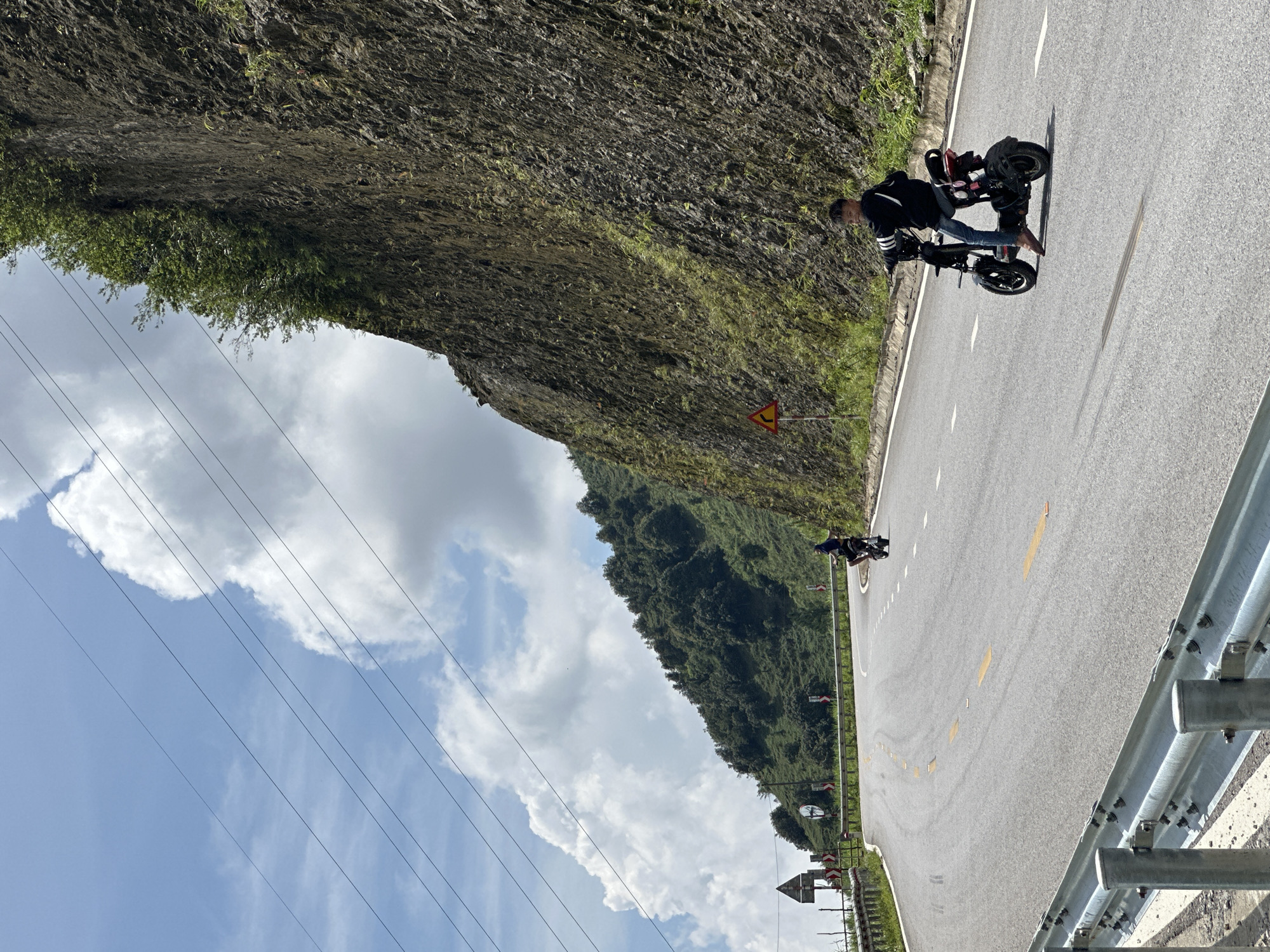 The image, taken in Muong Than, Vietnam, captures a sunny day on a winding mountain road. A paved road, marked with white dashed lines and yellow solid lines, curves through the landscape. To the left, a steep, dark rock face, partially covered with green foliage, dominates the view. Opposite, a lush, tree-covered hillside slopes downwards.
In the foreground, a man is standing beside a compact electric motorcycle or scooter. He wears a dark long-sleeved top with white stripes on the sleeves and blue jeans, appearing barefoot. He is looking towards the viewer, possibly taking a break from riding. Further down the road, another person on a similar vehicle is visible.
A silver metal guardrail runs along the right side of the road. Power lines stretch across the bright blue sky, which features scattered white clouds. A yellow triangular road sign with a black squiggly arrow, indicating a series of bends, is prominent near the rocky mountain face. Another sign and a traffic mirror can be seen further along the road. The strong sunlight casts clear shadows, suggesting it is midday or early afternoon, highlighting the scenic and rugged terrain.