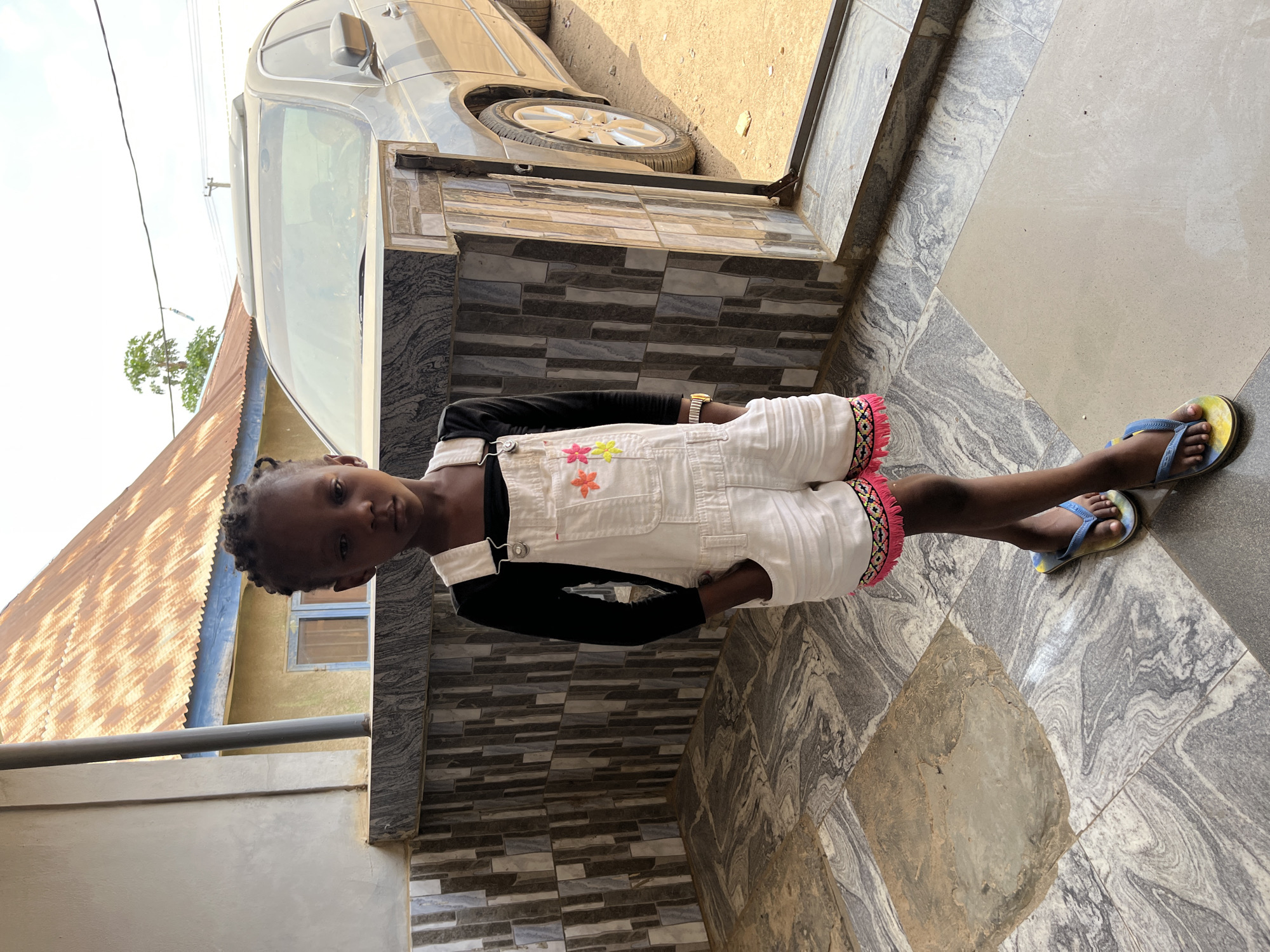 A girl stands on a tiled surface outside a building in Madalla, Nigeria. She wears a long-sleeved black shirt under white overalls with floral designs. Her footwear consists of blue and yellow flip-flops. A car is partially visible near the top of the frame, and a house with a corrugated metal roof can be seen in the background.