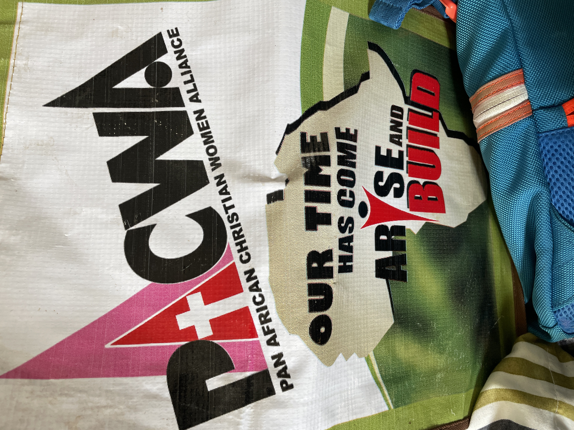 A partially visible white woven plastic bag with black text reading "PACWA PAN AFRICAN CHRISTIAN WOMEN ALLIANCE" is visible. A logo composed of a black "P", a pink triangle, and a red triangle with a white cross is also on the bag. A torn piece of paper with the text "OUR TIME HAS COME ARISE AND BUILD" lies on top of the bag, partially obscuring its text. The torn paper partially covers a map of Africa rendered in shades of green. Portions of a blue backpack and a striped cloth are also visible in the image. The city of Jos, Nigeria is not discernible from the provided image.