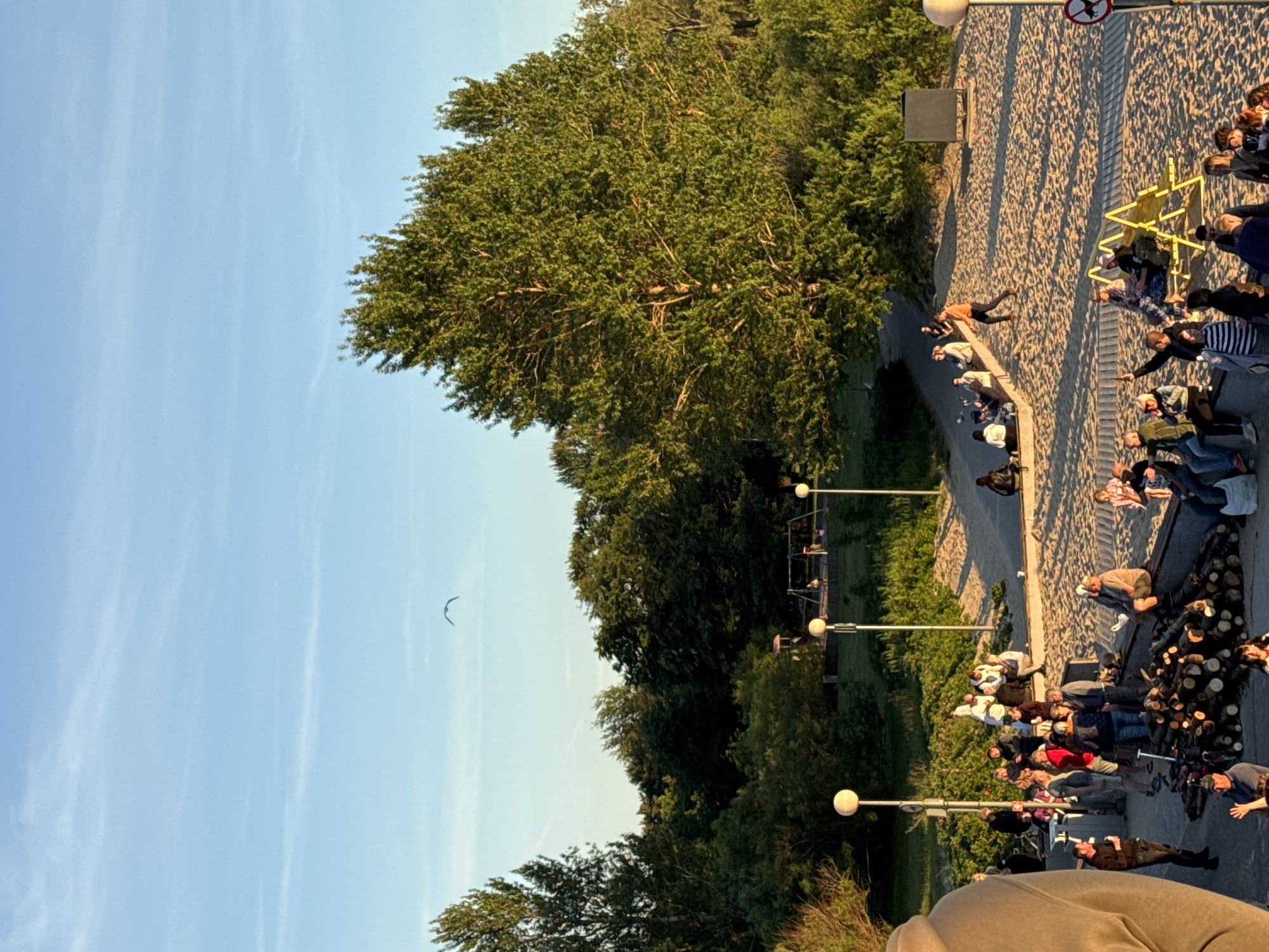 The image depicts a sunny outdoor scene on a sandy area, likely a beach or park, with a clear blue sky and some thin clouds. A flock of birds is flying in the distance. In the foreground, there is a large pile of cut logs, suggesting a potential bonfire or gathering. A group of people are gathered around the logs and on a low wall, some appearing to be interacting and others seated. Further in the background, a playground with swings can be seen, and more trees and greenery line the area. The lighting suggests it is late afternoon or early evening. The scene is set in the city of Tallinn, Estonia, specifically at the Pirita beach promenade area, a popular public space for recreation and social gatherings.