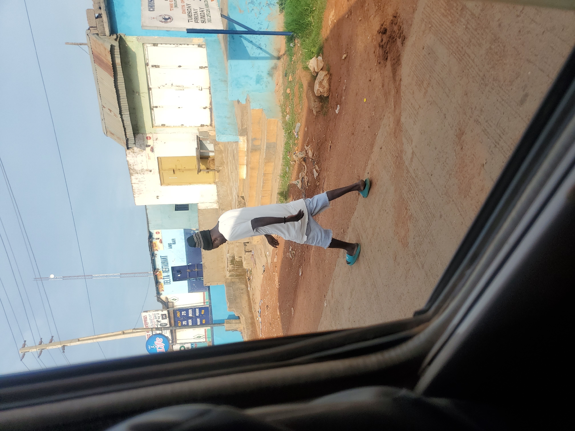 This is a photograph taken from inside a vehicle, looking out at a street scene in Goni Gara, Nigeria. The foreground is dominated by the dark interior of the vehicle's window frame. In the mid-ground, a person wearing a white t-shirt, light blue shorts, a dark beanie, and blue flip-flops is walking from left to right across the image. They are walking on a paved surface that appears to be a sidewalk or a part of the road. Behind the person, there are buildings with various paint colors and architectural styles, including some with corrugated metal roofs. One building has a sign that reads "DELTA RESTAURANT" and "AMB. BAR". Another sign, partially visible, displays text related to "CHRIST THE KING" and lists days of the week. The environment appears to be somewhat dusty and shows signs of wear and tear, with some debris scattered on the ground. Utility poles and wires are visible in the background, indicating the presence of infrastructure. The sky is clear and pale blue.