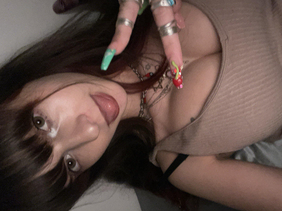 A woman with dark hair and bangs poses intimately. Her tongue is partially visible. She wears several rings on her fingers, adorned with long, decorated nails. A collection of necklaces hangs around her neck. She's dressed in a light brown top, revealing a portion of her chest. A black strap is visible on her arm. The photo has a close-up perspective and is taken at an angle.
