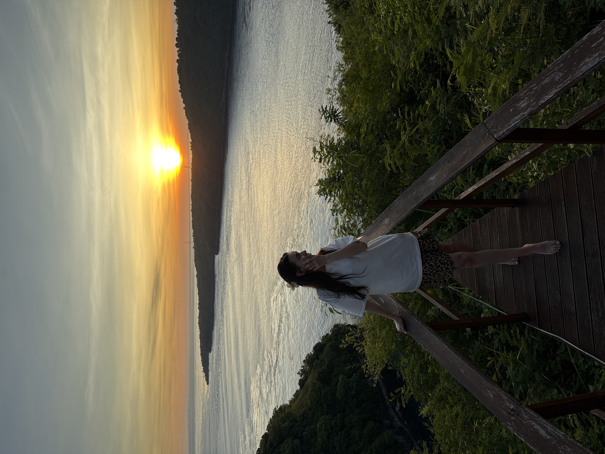 The image shows a woman standing on a wooden platform or boardwalk, possibly part of a viewpoint structure, overlooking a body of water and landscape at sunset. The sun is setting over what appears to be a coastline or a landmass across the water, casting a warm orange glow in the sky. The woman is wearing a white shirt and a patterned skirt or shorts. She is leaning against the railing and appears to be looking out at the view. The surrounding environment includes lush greenery and trees. The location may be somewhere in Nusa Penida, Indonesia, based on the environment.