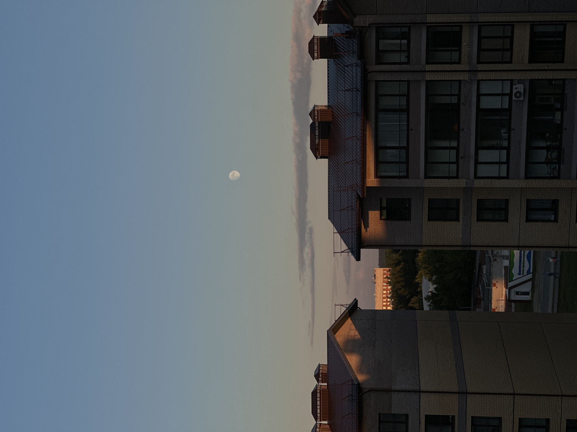The image shows a view of buildings with a sky background. The image is oriented vertically. In the sky, a full moon is visible. The buildings appear to be apartment blocks, constructed with light-colored bricks and dark-colored roofs. Some buildings have multiple windows and balconies. The setting suggests an urban or suburban environment.
The lighting indicates that it is either dawn or dusk, with the warm glow of the setting or rising sun casting shadows on the buildings. The sky is a gradient of blue and pale orange, with some wispy clouds. The overall mood is calm and serene. There are no people or active events clearly depicted in the image. The visible text in the image is a sign on a small building in the distance, which appears to be in Russian. The text reads "Нефтепромстрой".