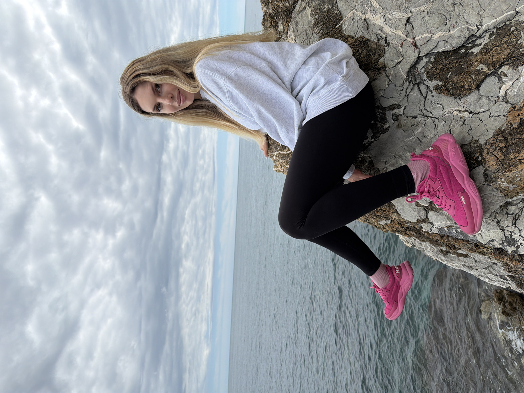 A woman is posed on a rocky cliff overlooking the sea. She has long blonde hair, is wearing a light gray sweatshirt, black leggings, and bright pink sneakers with matching socks. The rocky cliff edge is gray and textured. Behind her is a vista of the sea under an overcast sky. The location is likely near Budva, Montenegro, given the coastal geography and visual style.