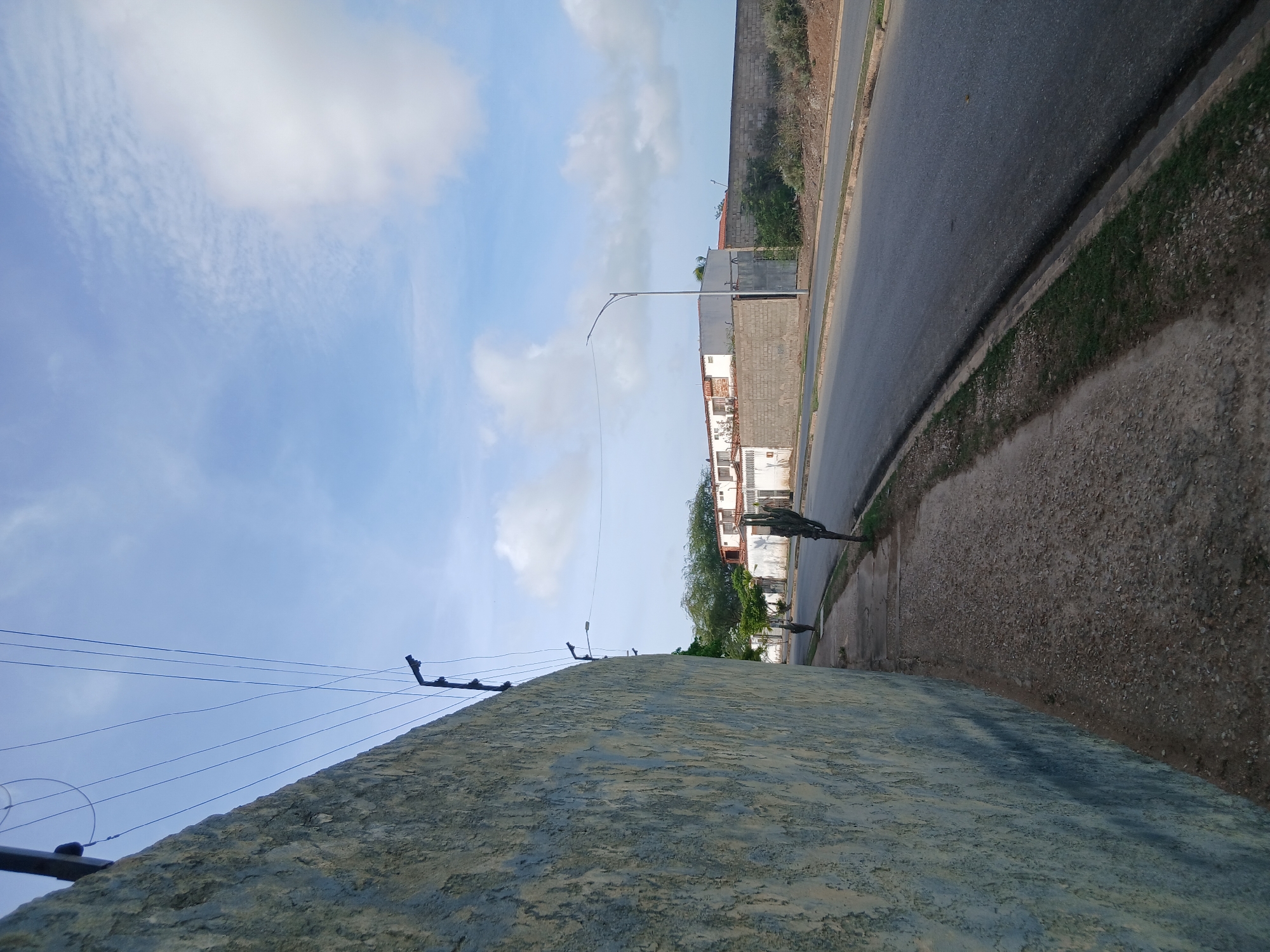 A street scene in Juan Griego, Venezuela, features a textured, light blue and yellow painted wall on the left foreground, extending towards the background. Above the wall, a series of thin, dark wires are strung diagonally across a pale blue sky with scattered white clouds. To the right of the wall, a sidewalk with a gravel and dirt surface runs parallel to an asphalt road. Along the sidewalk, two tall, thin cacti are visible. In the middle ground, buildings with white walls and red-tiled roofs are situated behind a grey brick wall. A single lamppost stands on the side of the road. The road itself is grey asphalt with a curb and a narrow strip of grass. A person is walking away from the camera on the sidewalk in the middle ground. The overall impression is of a somewhat arid, urban environment under a bright sky.