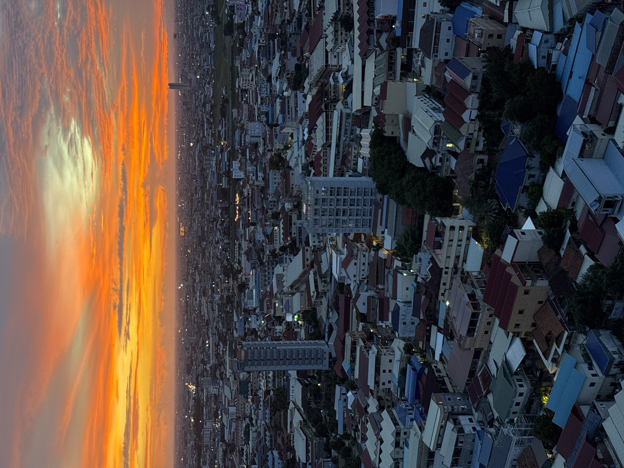 This is an aerial, vertical, eye-level, wide shot of a city at sunset in Khan Mean Chey, Cambodia. The sky is filled with vibrant orange and yellow clouds that are fanning out from the left side of the frame, creating a dramatic sunset. Below the sky, the city stretches out to the horizon, a dense collection of buildings with a variety of roof colors, including red, blue, and white. There are several tall buildings scattered throughout the cityscape, and some of them have lights on, indicating that it is evening. The overall impression is one of a bustling urban environment bathed in the warm glow of a setting sun.