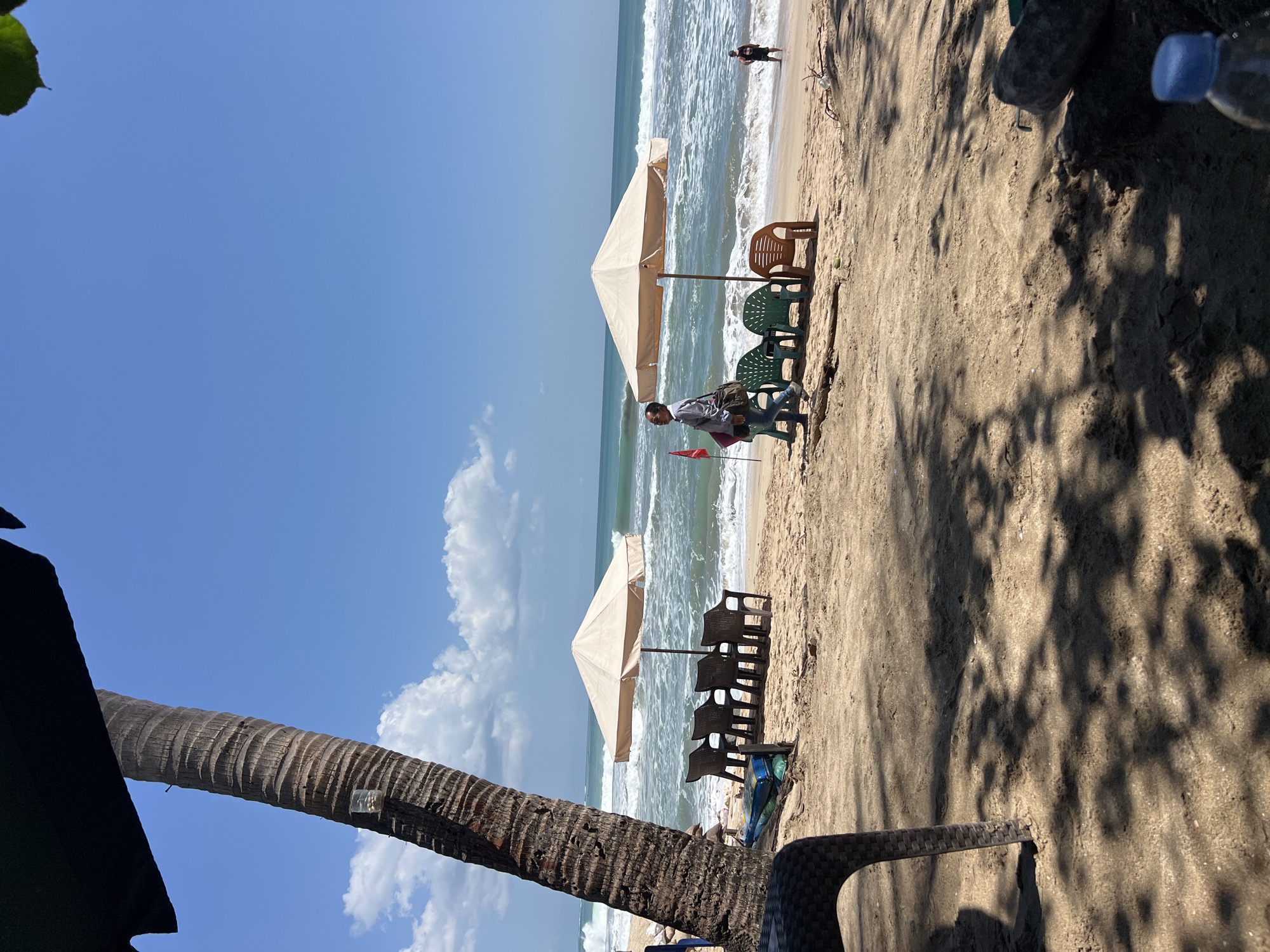 An eye-level, medium shot captures a sunny day at Kuta Beach in Bali, Indonesia. The foreground is dominated by the sandy beach and the textured leg of a dark chair, partially obscured by dappled shadows from an unseen tree or umbrella. To the left, a large palm tree trunk, its bark deeply grooved, rises towards the bright blue sky dotted with fluffy white clouds.
In the mid-ground, a row of dark plastic chairs sits on the sand, facing the ocean, with a light beige umbrella providing shade over the leftmost chairs. Further to the right, another beige umbrella shades three green plastic chairs and one brown plastic chair. Two people are visible on the beach. On the left, a person in a light-colored shirt and jeans walks away from the viewer, carrying a bag over their shoulder. On the right, near the water's edge, a second person stands facing the ocean, wearing dark clothing.
The ocean stretches across the background, with white-capped waves breaking on the shore under a clear blue sky. The overall impression is one of a serene beach day in a popular tourist destination.