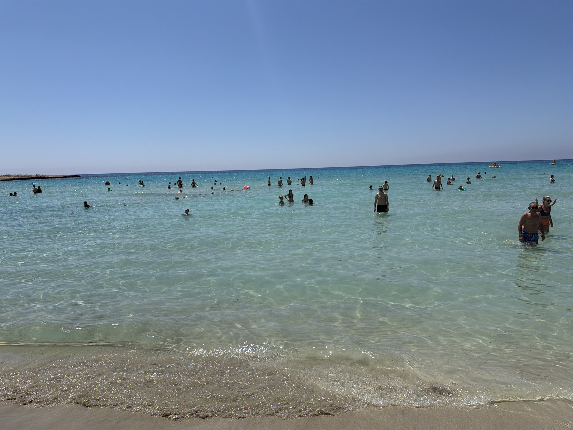 The image shows a beach in Ayia Napa, Cyprus, with numerous people wading and swimming in clear, turquoise water. The sky is clear blue. The foreground features the sandy shoreline with visible wave action. The background includes a rocky landmass. The people are diverse in age and attire. The water appears shallow, with the seabed visible in certain areas.