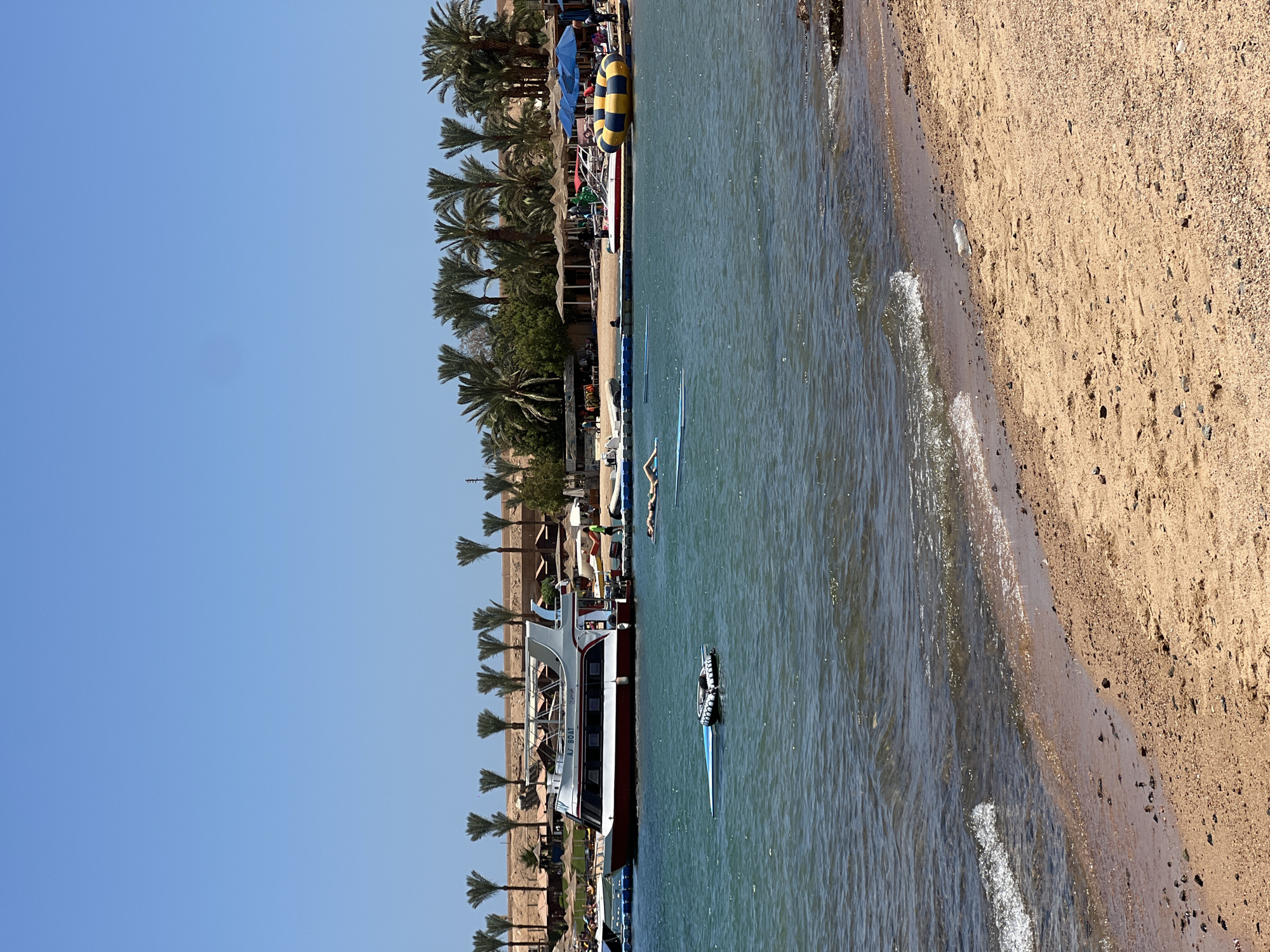 This image depicts a sunny day at a beach in Sharm El Sheikh, Egypt. In the foreground, a sandy beach meets the clear blue water, with small waves gently lapping at the shore. A partially submerged object, possibly a damaged inflatable or a small boat, is visible in the water. Further out, a large white and red boat with the text "AJ BOAT" is docked. Several people are present, with one person seen relaxing on an inflatable mat in the water and others walking on a blue pontoon walkway that extends into the sea. In the background, palm trees line the shore, behind which are buildings and beach umbrellas. The sky is a clear, bright blue.