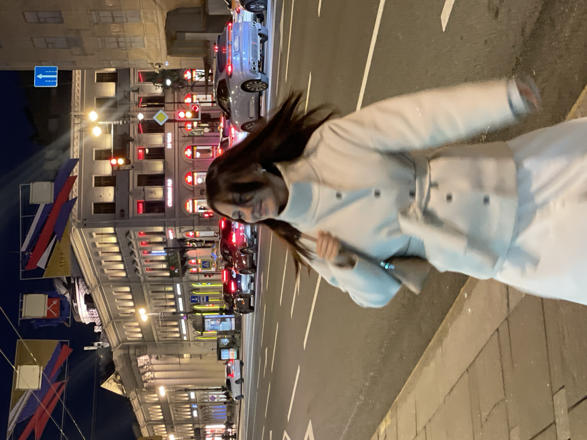 A woman is walking along a street in what appears to be Saint Petersburg, Russia, at night. The background features a row of buildings illuminated by streetlights, along with cars, a traffic light, and Russian flags hanging from lampposts. The woman is wearing a light-colored outfit with a belt. The image is slightly blurry, likely due to movement.