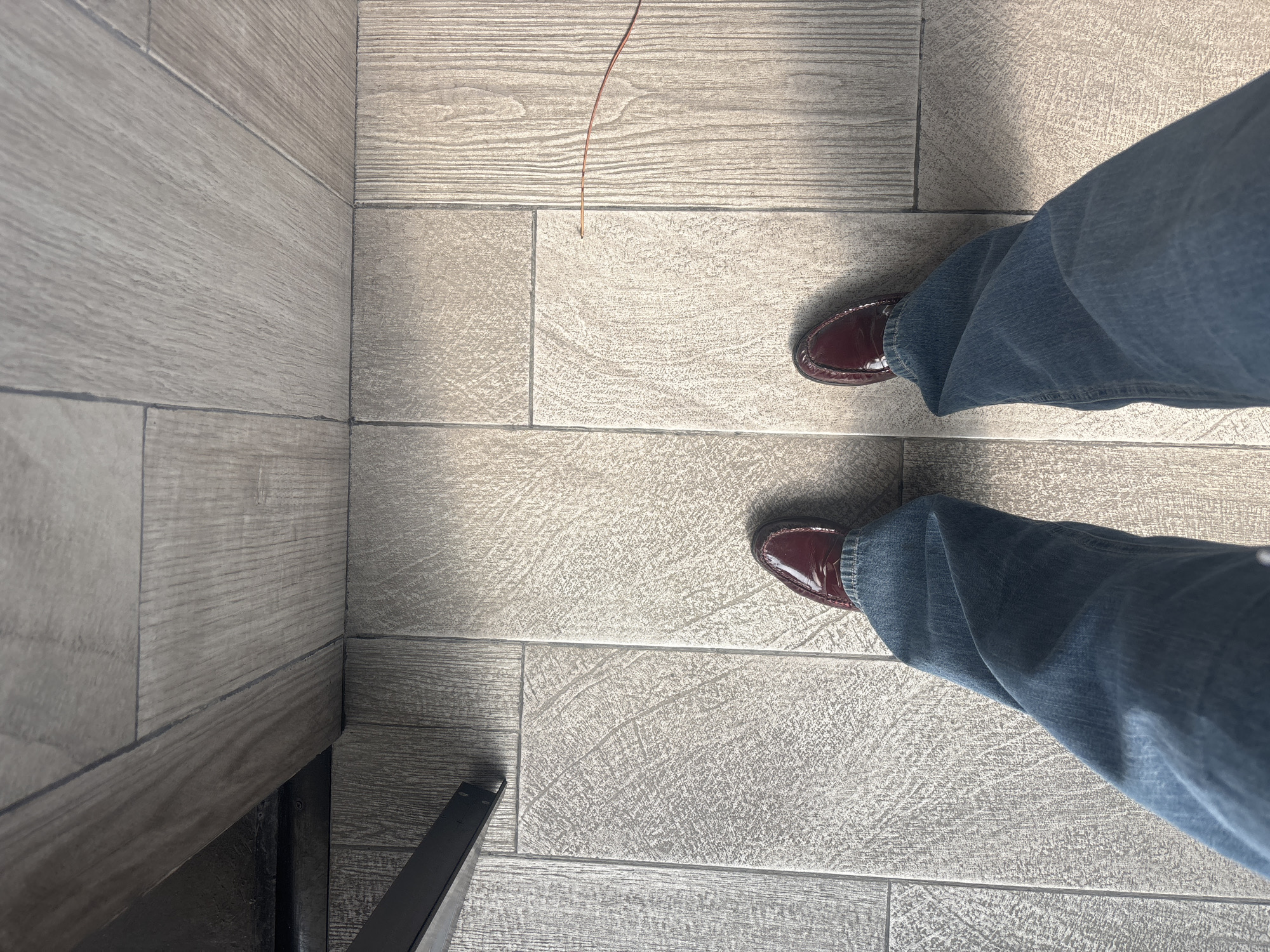The image shows a top-down view of a person's lower body, wearing blue jeans and burgundy loafers, standing on a tiled floor. The floor consists of rectangular tiles with a textured, stone-like pattern. A section of wooden-look flooring with linear grain is visible on the left side of the image. A thin, brown object, possibly a piece of straw or thread, is present on the floor near the top center of the frame. The lower left corner contains a dark, indistinct object, possibly furniture legs. The location is unknown.
