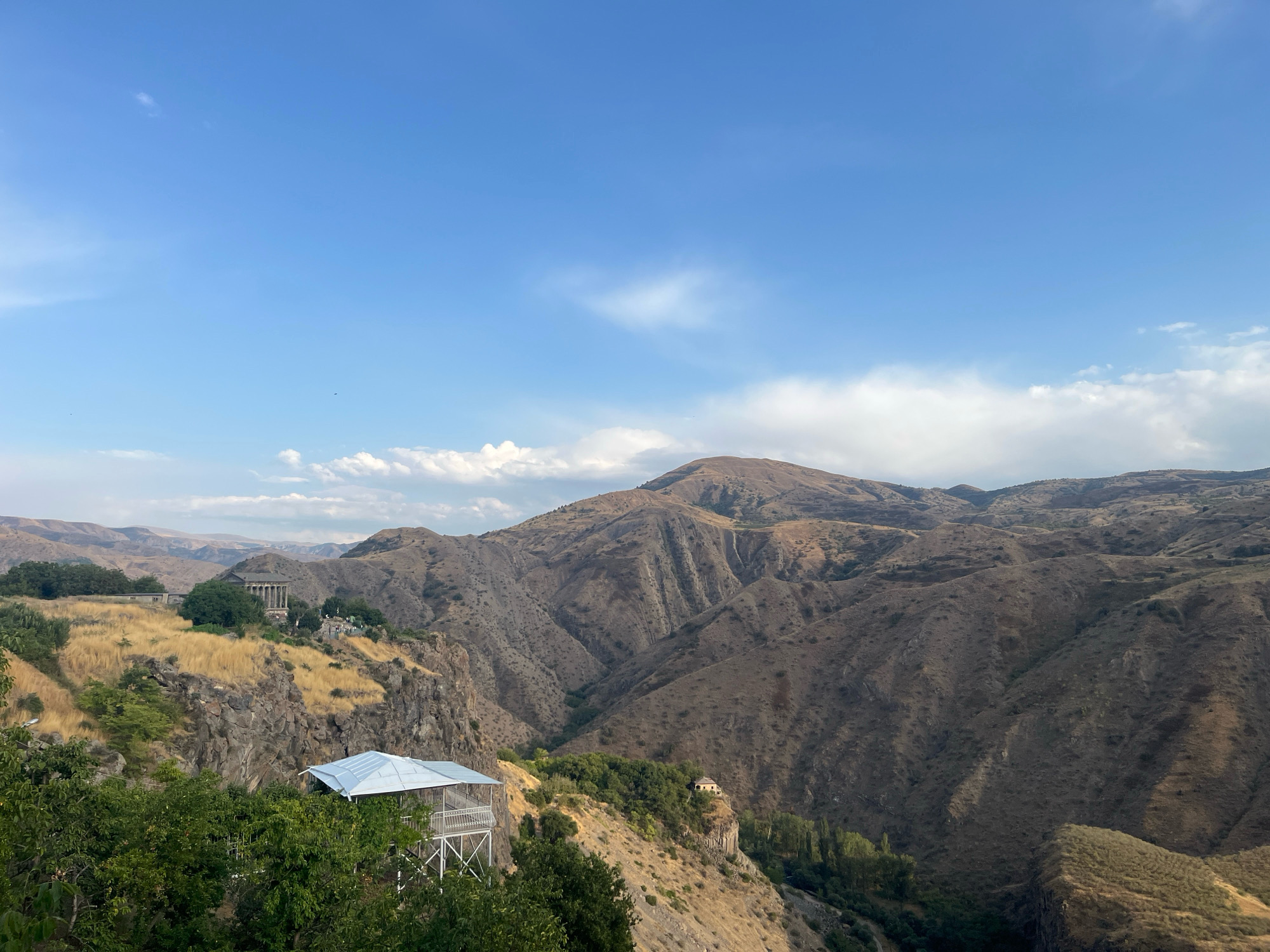 A wide-angle, landscape photograph captures a view of the Garni Gorge and the surrounding mountainous terrain in Garni, Armenia. The sky is a clear blue with scattered white clouds, particularly prominent on the right side of the frame. In the foreground, lush green trees occupy the lower left portion of the image.
Dominating the middle ground is a white, open-sided pavilion with a silver roof, supported by a white metal framework. It is perched precariously on the edge of a steep, rocky cliff face. The cliff itself is rugged and textured, with visible striations and patches of dry grass.
Further back, across the gorge, the landscape is characterized by rolling, arid mountains and hills. The slopes are covered in dry, brown vegetation, with deep ravines and valleys carved into the terrain, creating dramatic shadows. On the far ridge, atop a hill, sits a classical-style structure with columns, likely part of the Garni Temple complex. Another smaller, isolated building with a light-colored roof is visible on a rocky promontory further into the gorge. The overall impression is one of rugged natural beauty and ancient architecture integrated into the landscape. There are no visible people in the frame.