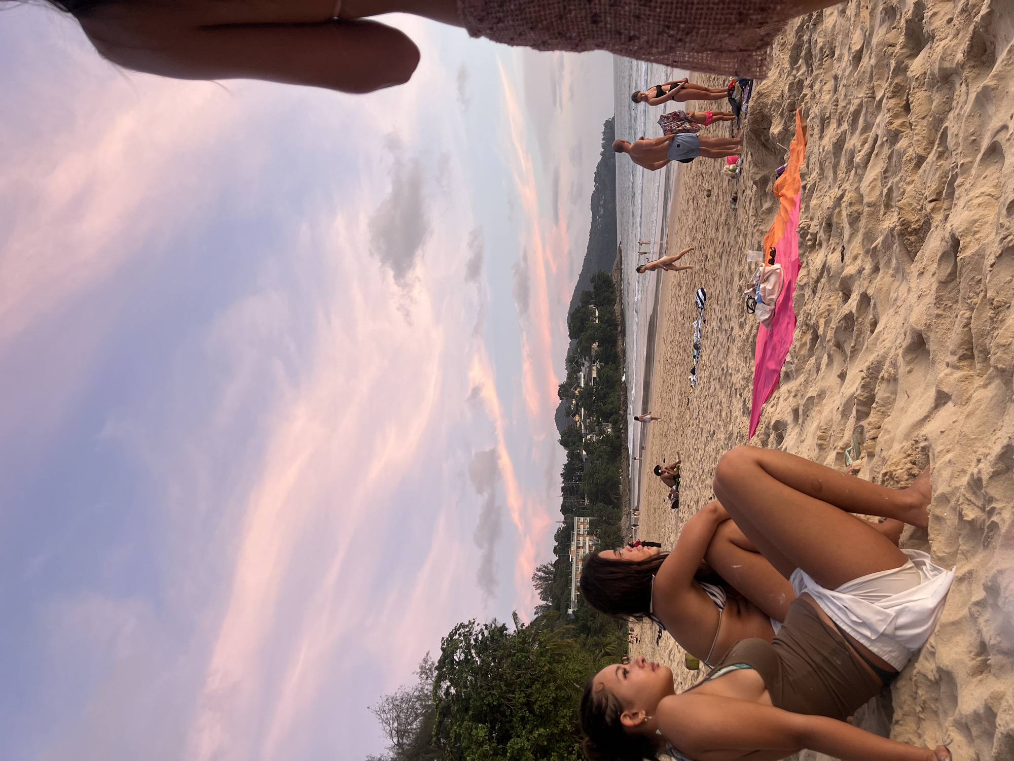 The image depicts a beach scene at dusk, with the sky painted in soft hues of pink and purple from the setting sun. Several people are visible on the sandy shore. In the foreground, two young women are seated, looking towards the water. One is wearing a striped bikini top and white shorts, while the other is in a brown bikini top and shorts.
Further down the beach, a man in blue shorts and a woman in a black bikini are standing near the water's edge. Another man is also visible in the distance, walking along the shoreline. A few colorful towels and beach bags are scattered on the sand.
The background features a treeline and buildings, suggesting a coastal town or resort area. The overall atmosphere is relaxed and tranquil, characteristic of a peaceful evening at the beach. There is no visible text in the image.