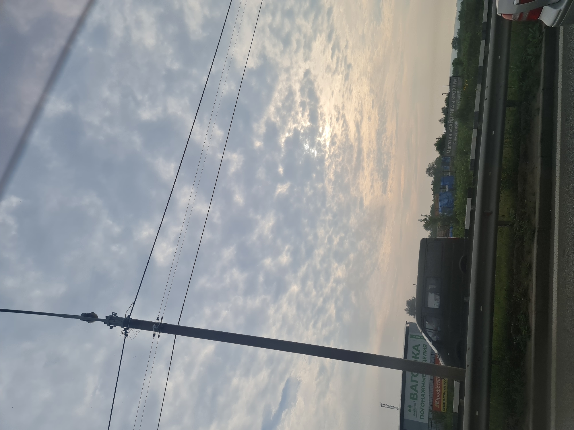 The image captures a cloudy sky with the sun peeking through, casting a soft light. A tall, dark utility pole with wires is prominent in the left half of the frame. In the mid-ground, a dark-colored van is parked alongside a highway barrier. Behind the van, a billboard with Russian text, including "ВАГОН КА" and "ПОГОНАЖНЫЕ ИЗДЕЛИЯ," is visible. Further in the background, trees and more billboards are present. The scene suggests an industrial or commercial area adjacent to a road, likely on the outskirts of Irkutsk, Russia, given the Cyrillic script. The overall lighting and cloud formation indicate it is either early morning or late evening.