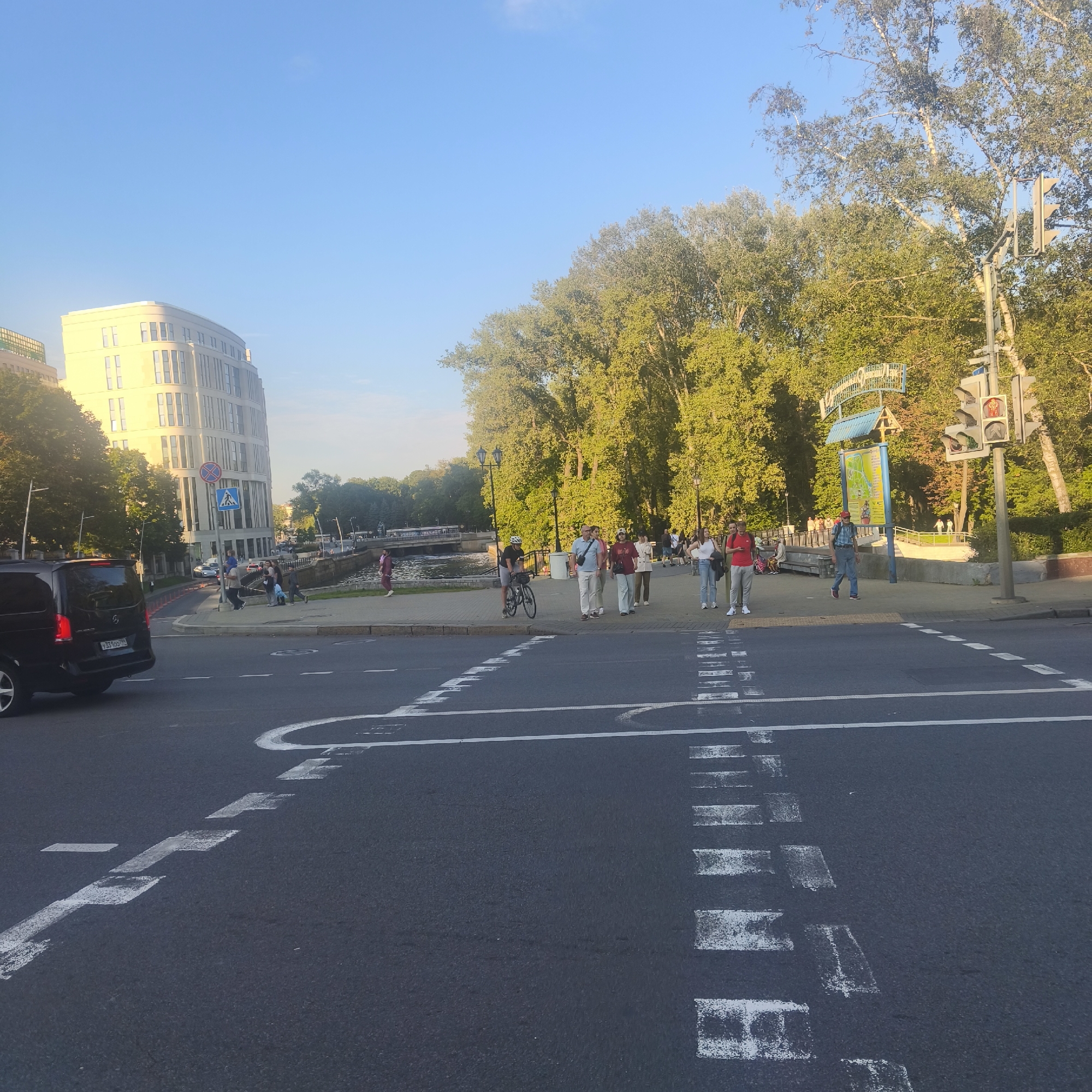 The image depicts an urban scene in Minsk, Belarus, likely near the Svislach River embankment, as suggested by the presence of a canal or river in the background and a distinctive building. In the foreground, a black Mercedes-Benz V-Class van is parked on the left side of the image, facing away from the camera, with its license plate visible. The street has white pedestrian crossing markings.
On the right side of the image, a group of people are walking and standing along a paved pedestrian walkway adjacent to the canal. Some individuals are carrying bags, and a cyclist is standing with his bicycle. A traffic light is visible, showing a red pedestrian signal. Behind the walkway, lush green trees form a canopy, and a modern, light-colored building with many windows is situated to the left. A blue sign with Cyrillic text and a map is mounted on a post near the traffic light. The sky is clear and blue, indicating daytime with bright sunlight. The scene appears to be a popular public area, possibly a park or promenade, in Minsk.