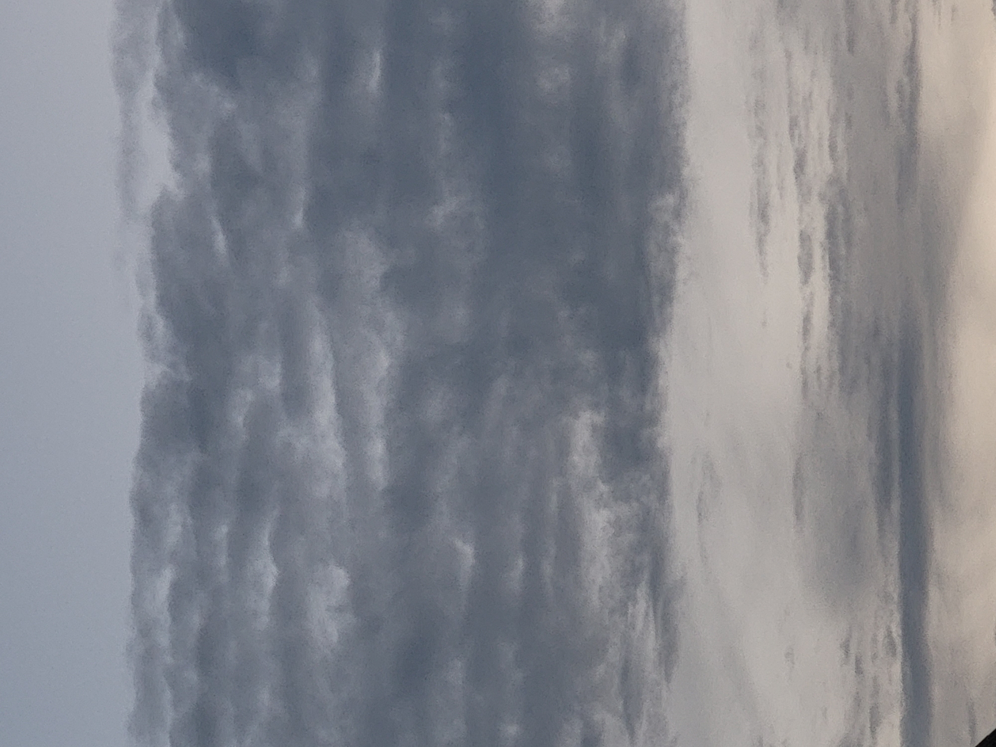 The image shows a cloudy sky at dusk or dawn. The clouds are gray and appear to be layered, with some lighter patches indicating breaks in the cloud cover. The sky takes up the majority of the frame, with a sliver of what appears to be land or buildings visible at the very bottom edge of the image. The overall lighting suggests a low light condition, possibly twilight. There are no people, specific landmarks, or identifiable objects in the scene to determine the location within a city.