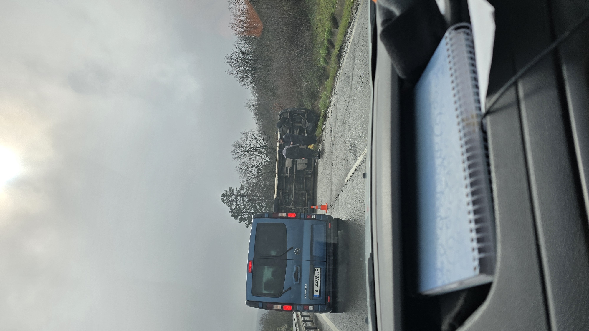 The image depicts a traffic incident on a highway, possibly near Burgas, Bulgaria, judging by the license plate on the blue Renault Vivaro van visible in the foreground. The van, with license plate "A 4498 HP," is positioned behind an overturned truck. Two people are standing near the overturned truck, seemingly examining the situation. An orange traffic cone is placed on the road near the van. The weather appears overcast and the environment includes trees and foliage lining the road. The picture appears to have been taken from inside a vehicle. A notebook is partially visible in the foreground of the image.