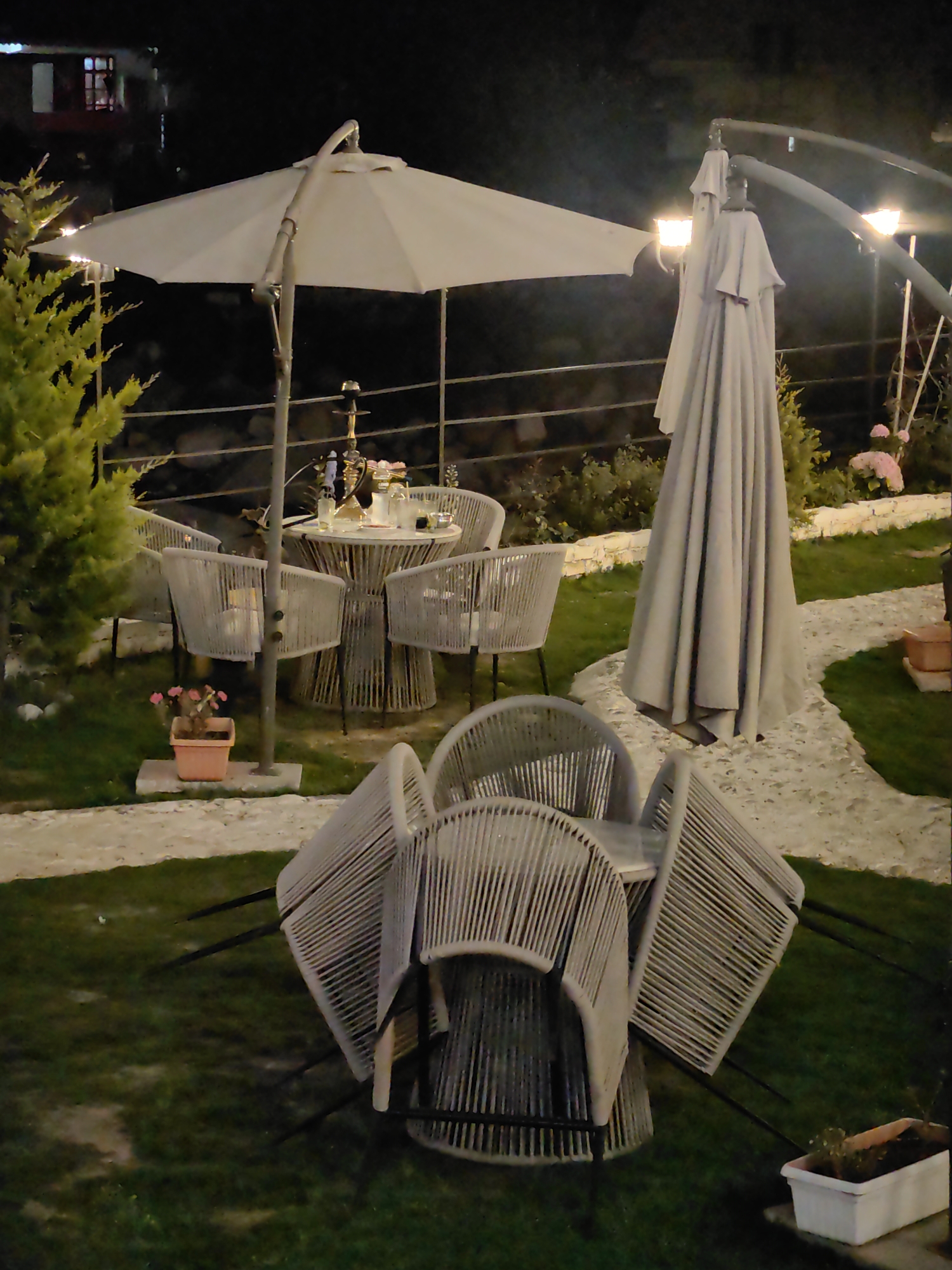 The image depicts an outdoor seating area at night, likely a cafe or restaurant, with a focus on tables and chairs. Several light-colored chairs, made of a woven material, are arranged around a round table. A large, closed umbrella is leaning against a pole in the center of the frame, and another opened umbrella is visible further back. Decorative plants and lighting fixtures are present in the background. The ground is a mix of grass and a paved walkway. The scene is set in a city environment, possibly Barheta, India, as suggested by the user, but this cannot be confirmed from the visual information alone. There are no people visible in the image.