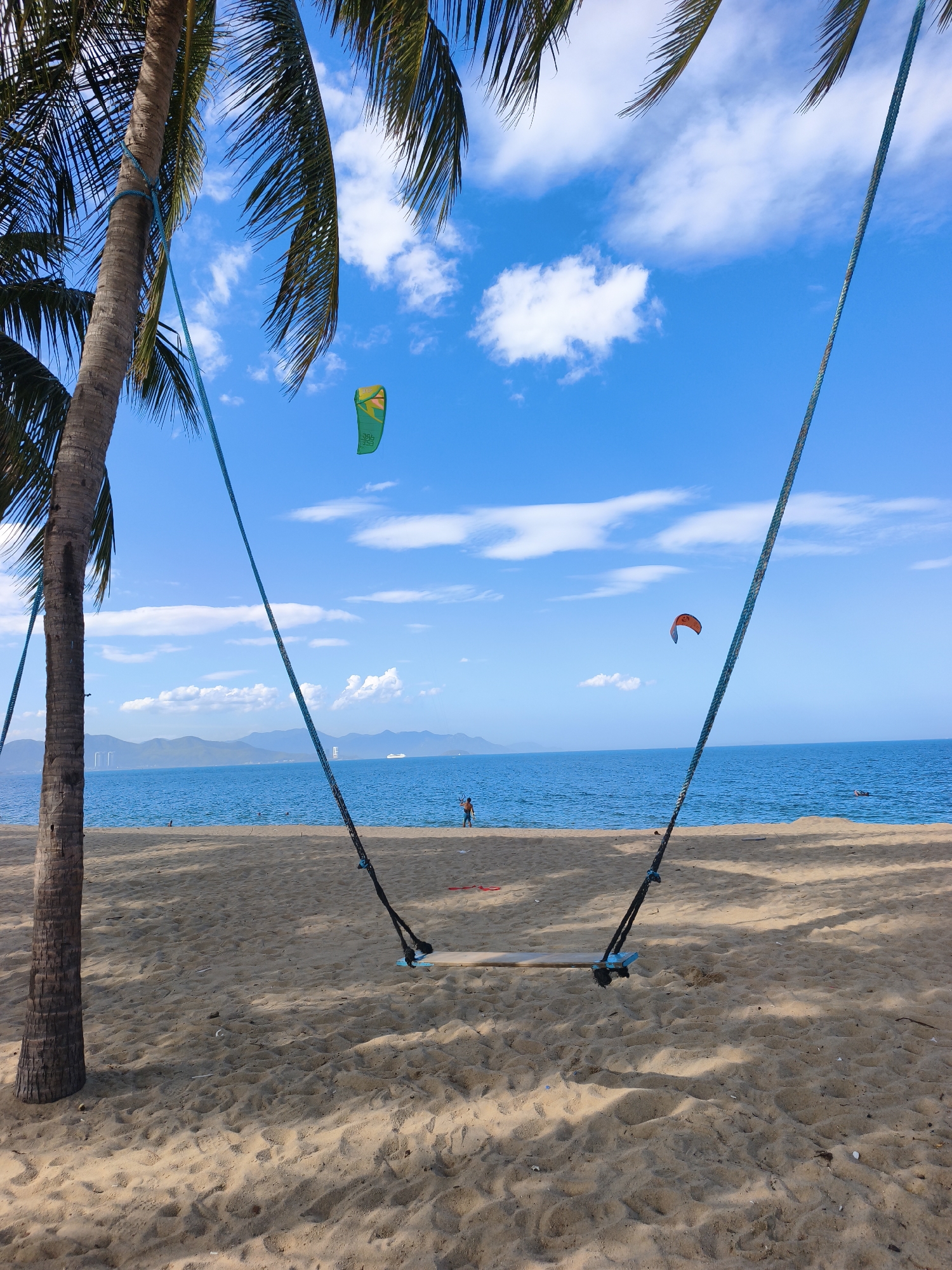 A swing hangs from palm trees on a sandy beach overlooking the ocean. The water is a deep blue, and the sky is a bright blue with scattered white clouds. In the distance, mountains can be seen on the horizon. There are several kitesurfers in the water. The scene is likely in Nha Trang, Vietnam, given the tropical setting and the presence of palm trees and kitesurfing.