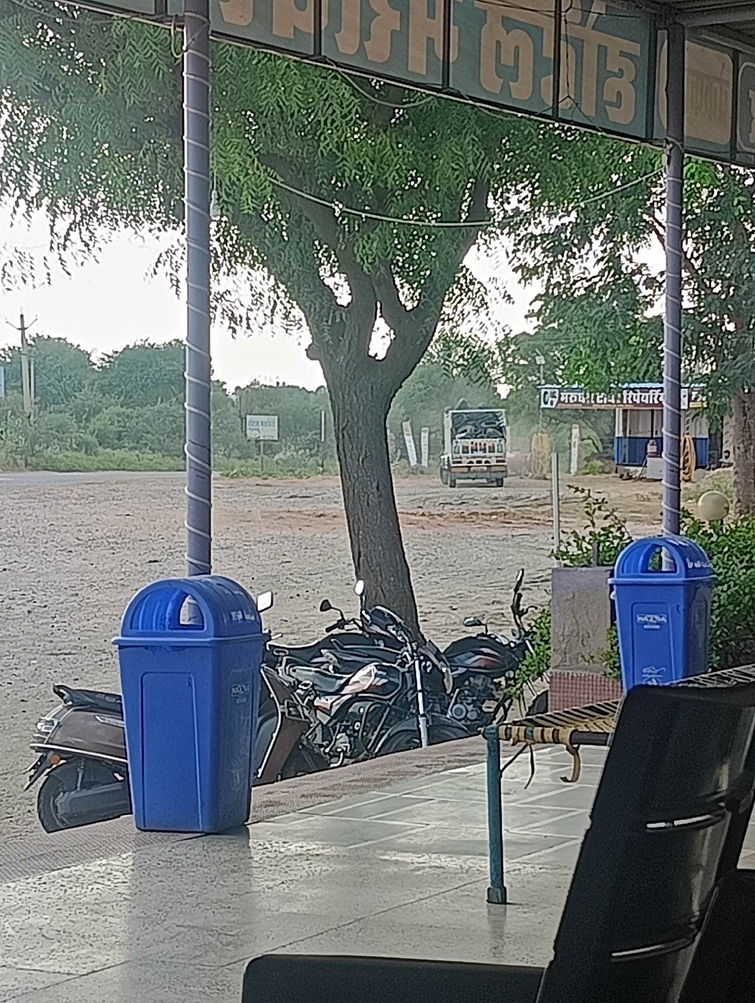The image depicts a street scene in Chhapri Bari, India, featuring a covered outdoor area with a tiled floor. In the foreground, two blue trash cans are positioned, with a scooter and two motorcycles parked to their left and right respectively. A truck is visible in the background, driving on a dirt road. To the right, a gas station with a blue and white structure is present. The scene is framed by trees and a sign with Hindi text.