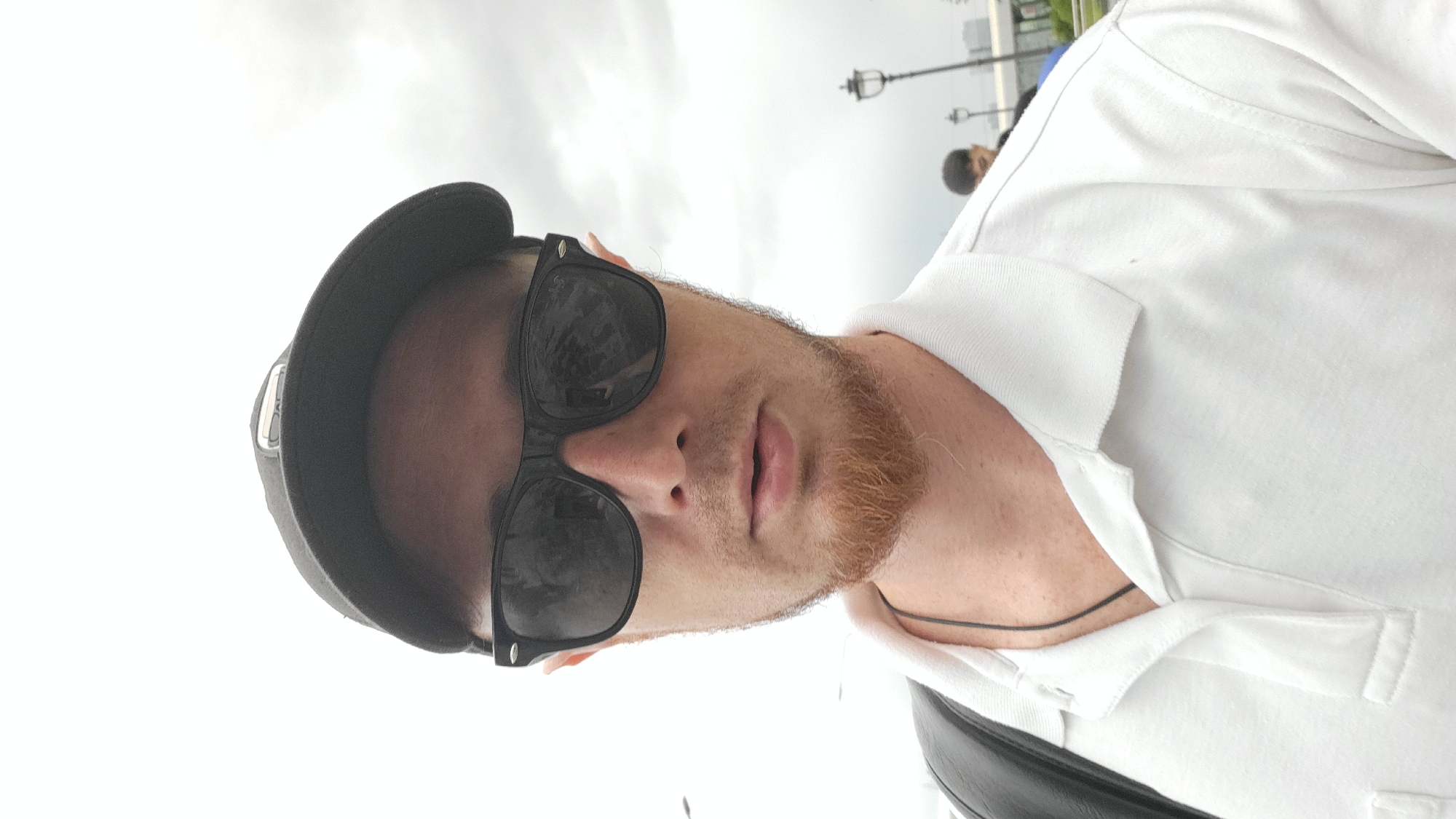 The media file is a portrait of a person wearing a white polo shirt and a black baseball cap. The person is also wearing black sunglasses, which obscure their eyes. They have a light complexion and a reddish-brown goatee. The image is taken from a low angle, looking up at the person. The background is bright and out of focus, suggesting an outdoor setting with a cloudy sky. There are some blurred figures and streetlights visible in the distance, hinting at an urban environment. The person's mouth is slightly open, giving the impression they might be speaking or about to speak. The lighting is diffuse, indicating it might be daytime, possibly overcast. There is a thin black string or cord visible around the person's neck, tucked into the polo shirt. The overall mood is casual and informal. No visible text is present in the image.