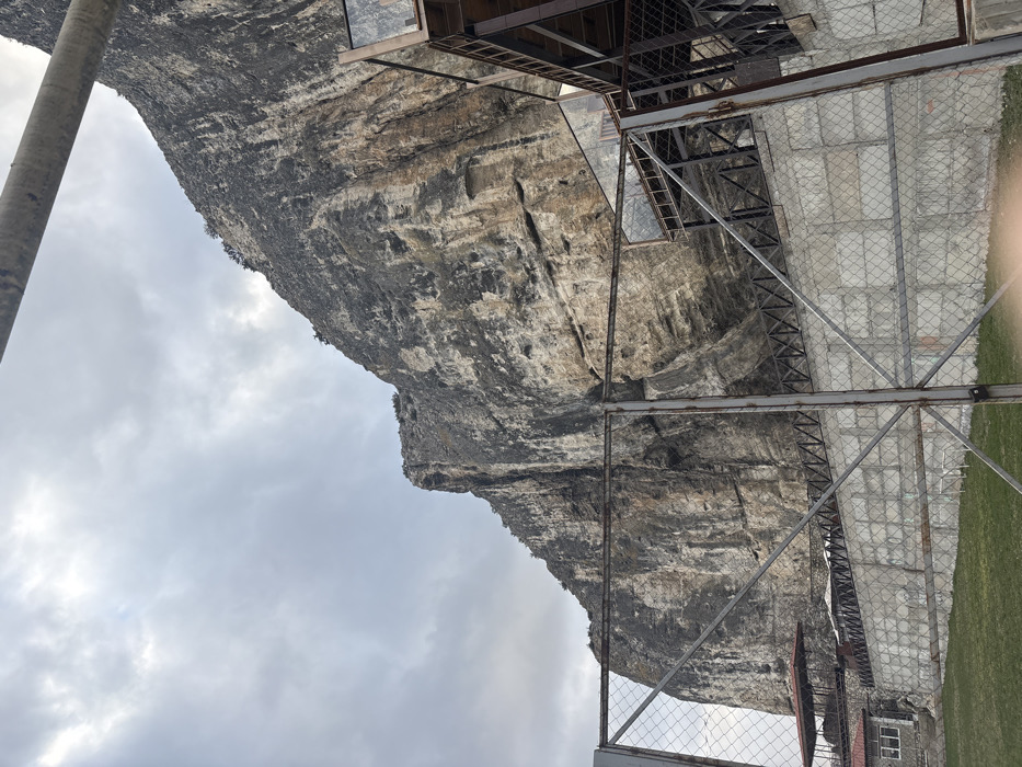 The image displays a prominent, rugged cliff face composed of varied grey, brown, and tan rock formations, rising steeply under a densely overcast, light grey sky. The scene is located in Bakhchysarai, Ukraine. In the foreground, a chain-link fence with vertical metal posts extends across the lower and middle sections of the frame. Behind this fence, a grey concrete retaining wall with a square block pattern is visible, topped by a dark metal railing. To the far left, behind the fence and wall, a building with a red-brown pitched roof and lighter-colored walls is partially visible, with another similar roofline further in the background. To the right, a multi-level structure constructed of dark metal framework and transparent glass panels is built into the base of the cliff, featuring visible stairs and platforms. A thick, light-colored cylindrical object or beam diagonally crosses the upper right corner of the frame, partially obscuring the cliff and sky. A patch of green grass is present in the lower-left corner of the foreground. No individuals or active interactions are depicted.
