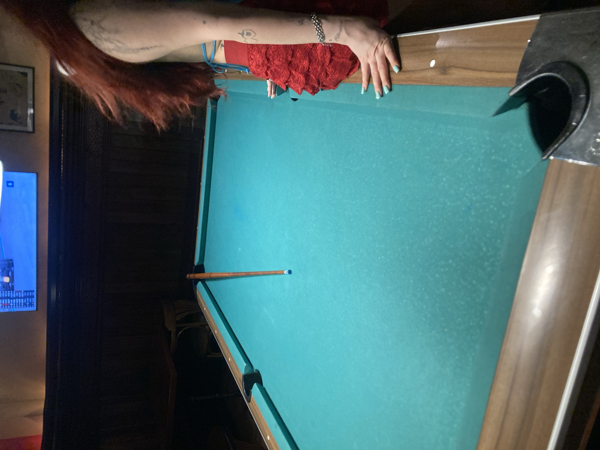 This image captures an indoor scene in Barcelona, Spain, within a dimly lit recreational area featuring a pool table. A person is partially visible on the right side of the frame, characterized by long, flowing reddish-brown hair and wearing a light blue top with a red, ruffled skirt. Their right arm, displaying multiple dark tattoos and a silver-colored beaded bracelet, rests on the wooden rail of the pool table, with light blue fingernail polish visible on their hand. A second hand, presumably theirs, is also placed on the table further along the rail.
The central object is a pool table with a teal-green felt playing surface, which exhibits a slightly dusty or worn appearance. The table features dark wooden rails with white circular sight markers. A single, light brown wooden pool cue with a blue tip lies horizontally across the table's felt surface. A dark corner pocket, possibly plastic with a metallic trim, is prominent in the lower right foreground.
In the background, dark wooden paneled walls are visible. Mounted high on the wall is a bright blue television screen displaying white text or data. To the right of the screen, a framed picture or piece of art hangs, though its details are indistinct due to lighting and distance. Further to the left, a dark wooden table and a chair with a curved backrest are partially visible, suggesting other seating arrangements within the establishment. The overall environment conveys the ambiance of a bar or pub.