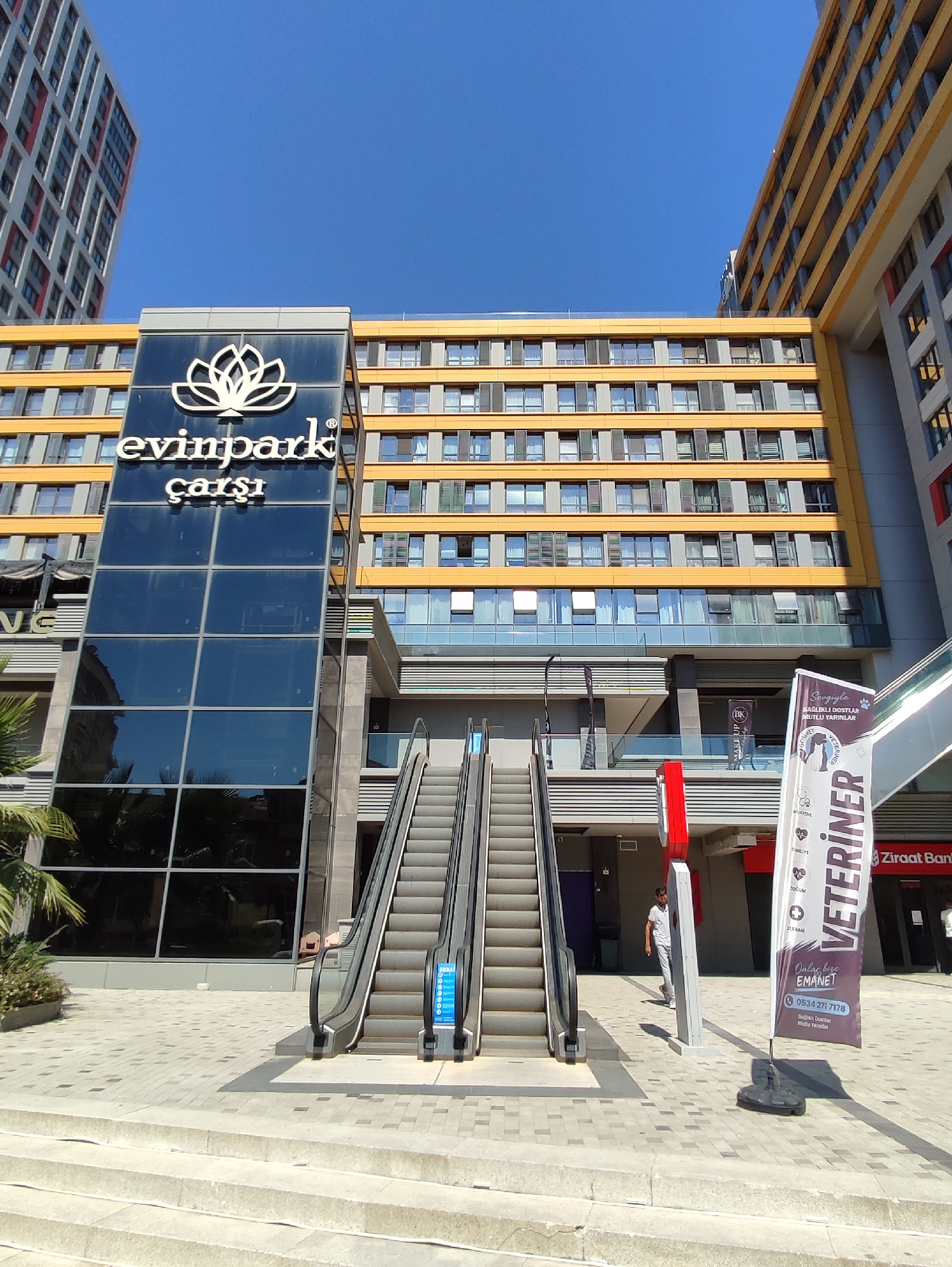 This is a medium-shot, eye-level, wide-angle outdoor photograph taken on a sunny day, facing a modern multi-story building complex with an attached glass structure. The building's facade is predominantly yellow with a grid of windows. A prominent glass-enclosed elevator shaft stands to the left, featuring a large white lotus flower logo and the text "evinpark çarşı" in white lettering. Two escalators ascend towards the building's entrance, with one in operation and the other stationary. To the right of the escalators, a tall, narrow banner displays the text "VETERİNER" vertically in large purple letters, along with smaller text below that includes a phone number. A man in a light-colored shirt and pants is walking away from the escalators towards the banner. Further to the right, a sign for "Ziraat Bankası" is visible. The ground is paved with interlocking grey and tan tiles, and concrete steps lead up to the plaza level. The sky is clear and blue. The scene appears to be in a commercial or mixed-use area in Kadıköy, Turkey, given the Turkish text on the signage.