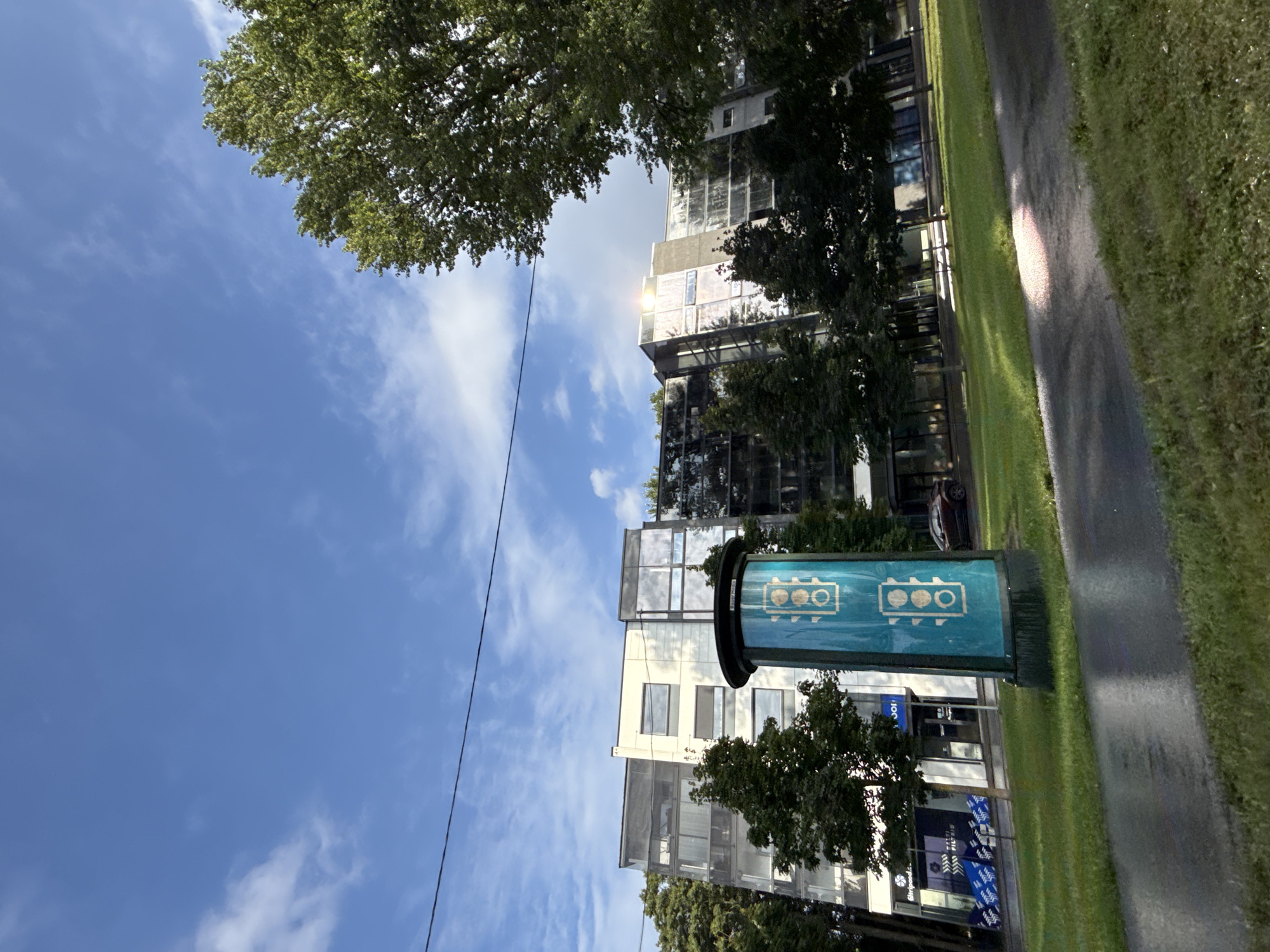 The image shows a modern, multi-story building with a glass facade and white and gray elements, set against a blue sky with scattered white clouds. In the foreground, there is a tall, cylindrical advertising pillar with a teal-colored background and white traffic light icons. To the left of the pillar, the building has large windows and a section with smaller, rectangular windows. Trees with lush green foliage are present in front of and around the building. A wet, paved walkway or road is visible in the lower part of the image, reflecting the sky and greenery. A dark-colored car is parked on the right side of the building. The overall scene suggests an urban environment in Tallinn, Estonia, possibly during daylight hours, with recent rain indicated by the wet surfaces.