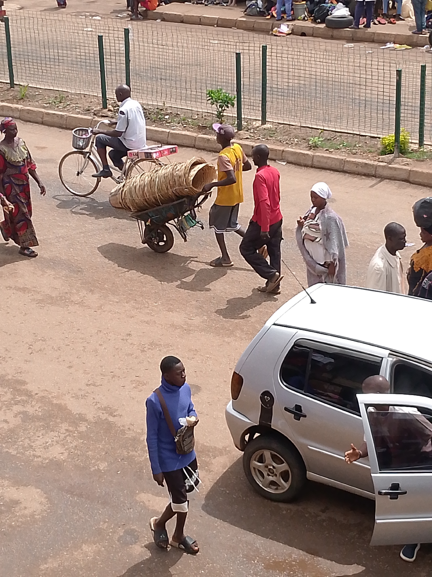 The image captures a bustling street scene in Bukuru, Nigeria. In the foreground, a young man in a blue turtleneck sweater and black shorts walks on a dirt road, carrying a small bag and a beverage. A silver Volkswagen Polo is parked on the right, with its front passenger door open, revealing a person inside. The street is lined with a metal fence, separating it from a raised curb with some sparse vegetation. Further in the background, various individuals are seen engaged in different activities. One man rides a bicycle laden with a basket of woven materials. Another person pushes a wheelbarrow also loaded with woven items. A woman in a colorful dress walks past, while a woman with a baby wrapped in a shawl stands nearby. Other people are visible in the distance, suggesting a lively marketplace or transportation hub. The overall atmosphere is one of everyday life and commerce in a Nigerian city.