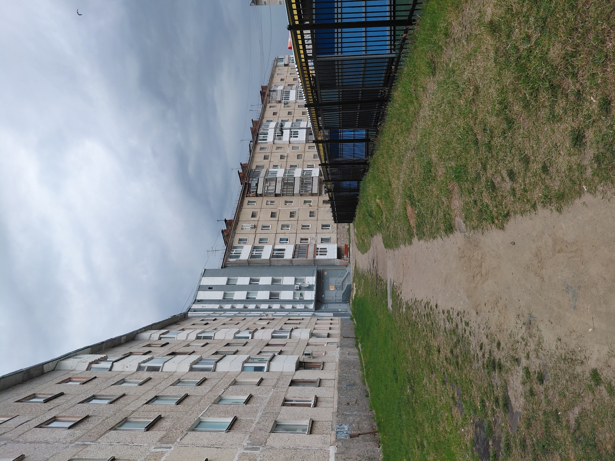 This image depicts a street in Nadym, Russia. On either side of a dirt path are Soviet-era apartment buildings with gray concrete exteriors and rows of rectangular windows. The buildings are multi-story. The sky is overcast. A black metal fence runs along the side of one of the buildings, separating it from a grassy area. The dirt path appears worn and compacted. A few antennae are visible on the roofs of the buildings.