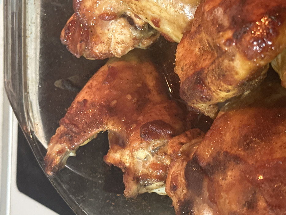 The image displays a close-up, overhead view of several pieces of cooked chicken in a clear glass baking dish. The chicken pieces appear to be wings or drumettes, golden brown and glistening with a rich, dark sauce or pan juices. Their skin shows a crispy texture and some areas have a slightly charred appearance, suggesting they have been roasted or baked.
The chicken is bathed in a considerable amount of glossy, dark brown liquid at the bottom of the dish. The individual pieces are arranged closely together, overlapping slightly. Details like the texture of the chicken skin and the reflections on the sauce are clearly visible due to the close-up framing.
The setting appears to be indoors, likely a kitchen, but no specific features of the room or background are discernible other than the clear glass of the baking dish. There are no people visible, and no activity beyond the static presentation of the cooked food. The lighting is bright, highlighting the sheen on the chicken and sauce. There is no visible text in the image.