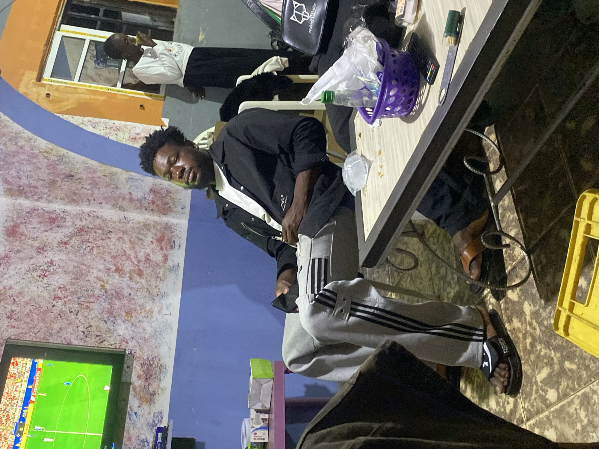 The image depicts an indoor scene, possibly in Jos, Nigeria, where a group of people are gathered. In the foreground, a man is seated in a chair, wearing a black jacket over a white t-shirt and grey sweatpants with black stripes. He is looking towards the camera. To his right, a small table is visible with a purple basket, a plastic bottle, and a remote control. In the background, another person is standing, wearing a white long-sleeved shirt and dark pants, holding an object to their mouth. A television screen displaying a soccer game is mounted on the wall on the left. The walls have a textured, multi-colored finish. The overall setting appears to be a casual, domestic environment.