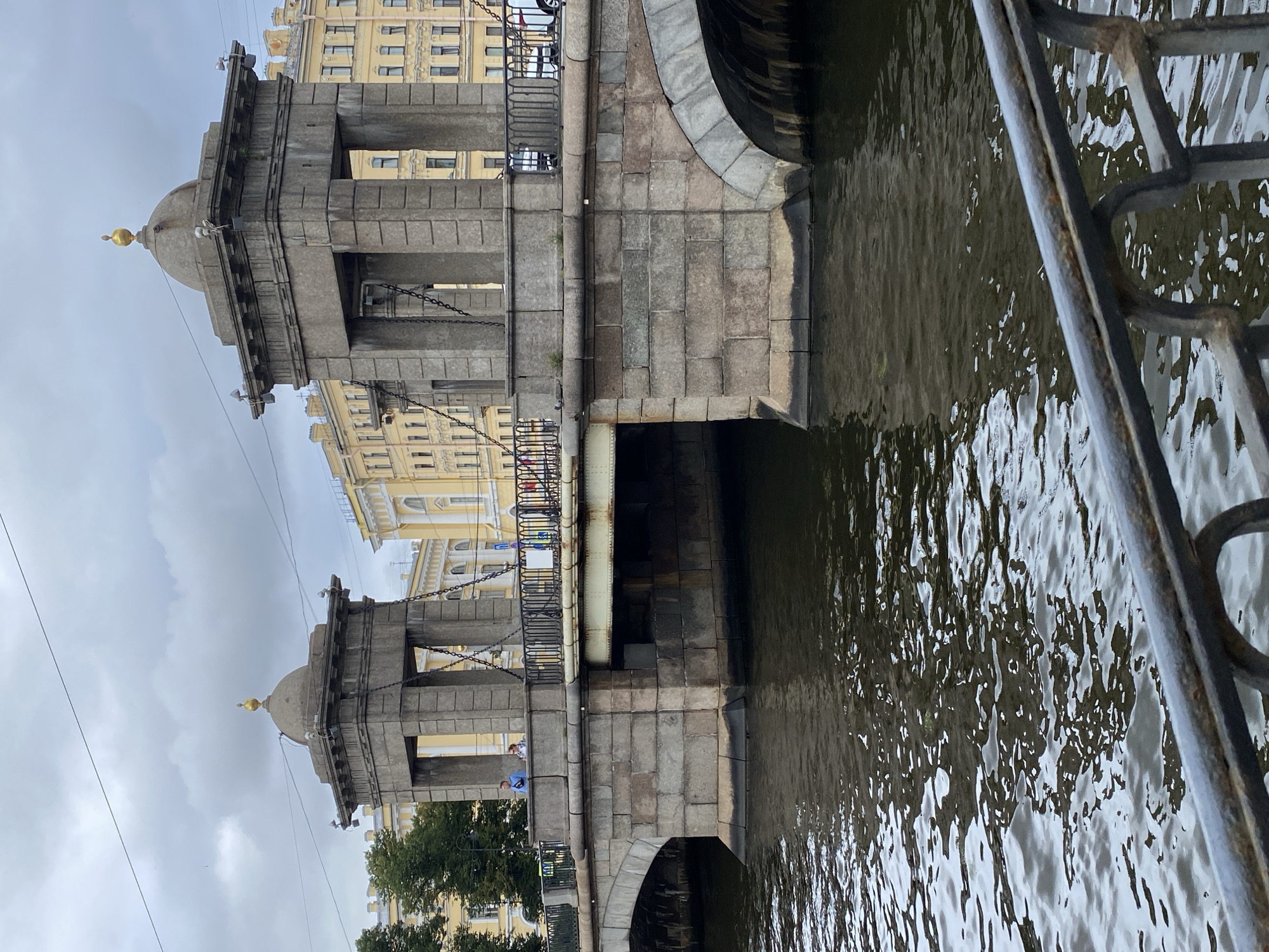 The image shows a view of the Bank Bridge over the Griboyedov Canal in Saint Petersburg, Russia. The stone bridge features two classical pavilions, each topped with a golden sphere. The canal water is dark and choppy, reflecting the overcast sky and the bridge's stone structure. In the background, a yellow building with many windows lines the street. A black metal railing is visible in the foreground, partially obscuring the view of the water. Two people are standing on the bridge, looking out. The scene depicts a typical cloudy day in Saint Petersburg.