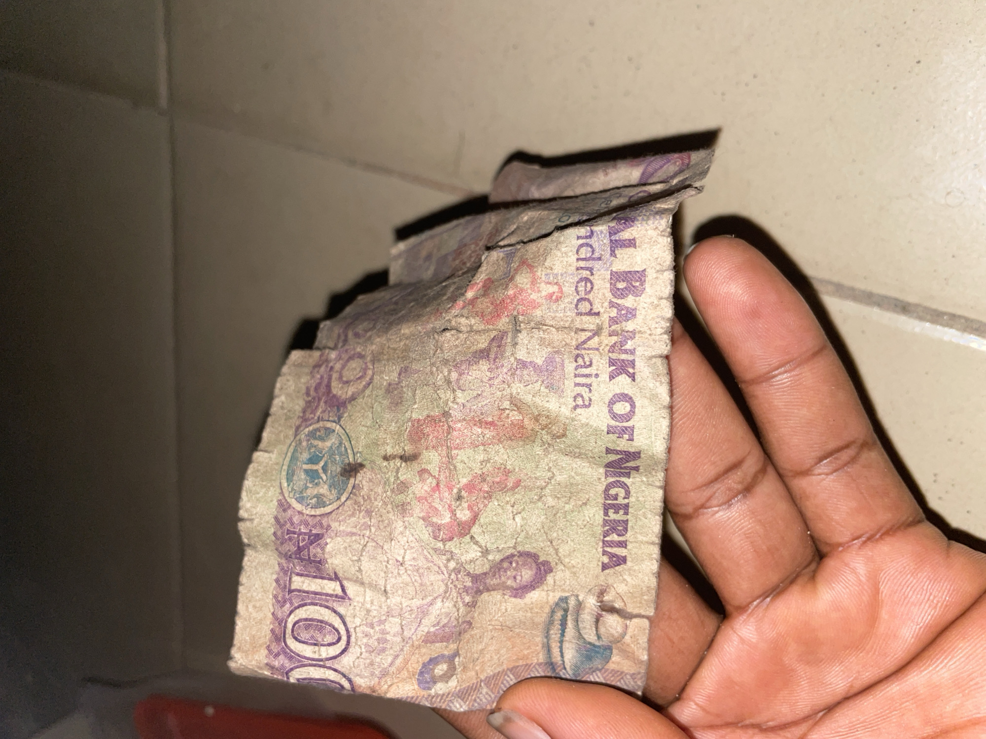 A hand holds a crumpled and torn Nigerian 100 Naira banknote. The note is faded and appears to be made of paper. The denomination "100" is visible in purple lettering on the left side of the note. Further text, partially obscured by creases, reads "BANK OF NIGERIA" and "hundred Naira" in purple ink. The background consists of a tiled floor and a dark, out-of-focus area. The image was taken in Jalingo, Nigeria.