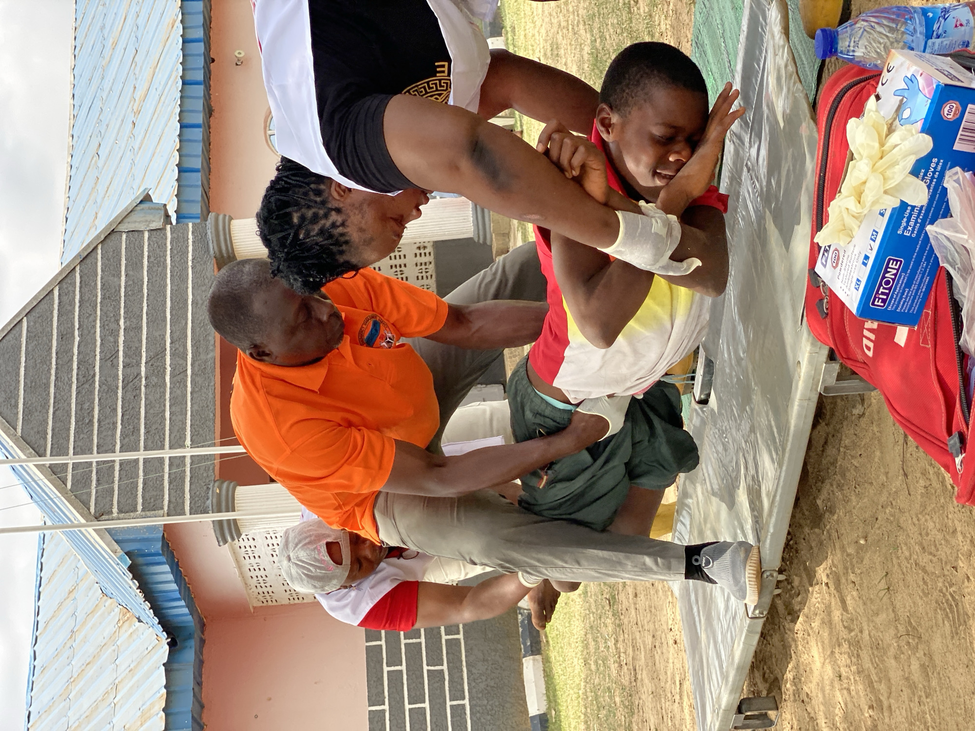 The image depicts a first aid scene. A young man in a yellow, white, and red shirt is being supported by three adults. One person, wearing a white and black top, supports his upper body. Another person, wearing an orange shirt, supports his torso. A third individual, wearing a white and red shirt and a white cap, supports his legs. The young man has a white bandage on his left arm. Medical supplies, including a box of Fitone examination gloves and a red first aid kit, are nearby. The scene appears to be outdoors, possibly in Nassarawa, Nigeria. A building with a blue and white roof is visible in the background.