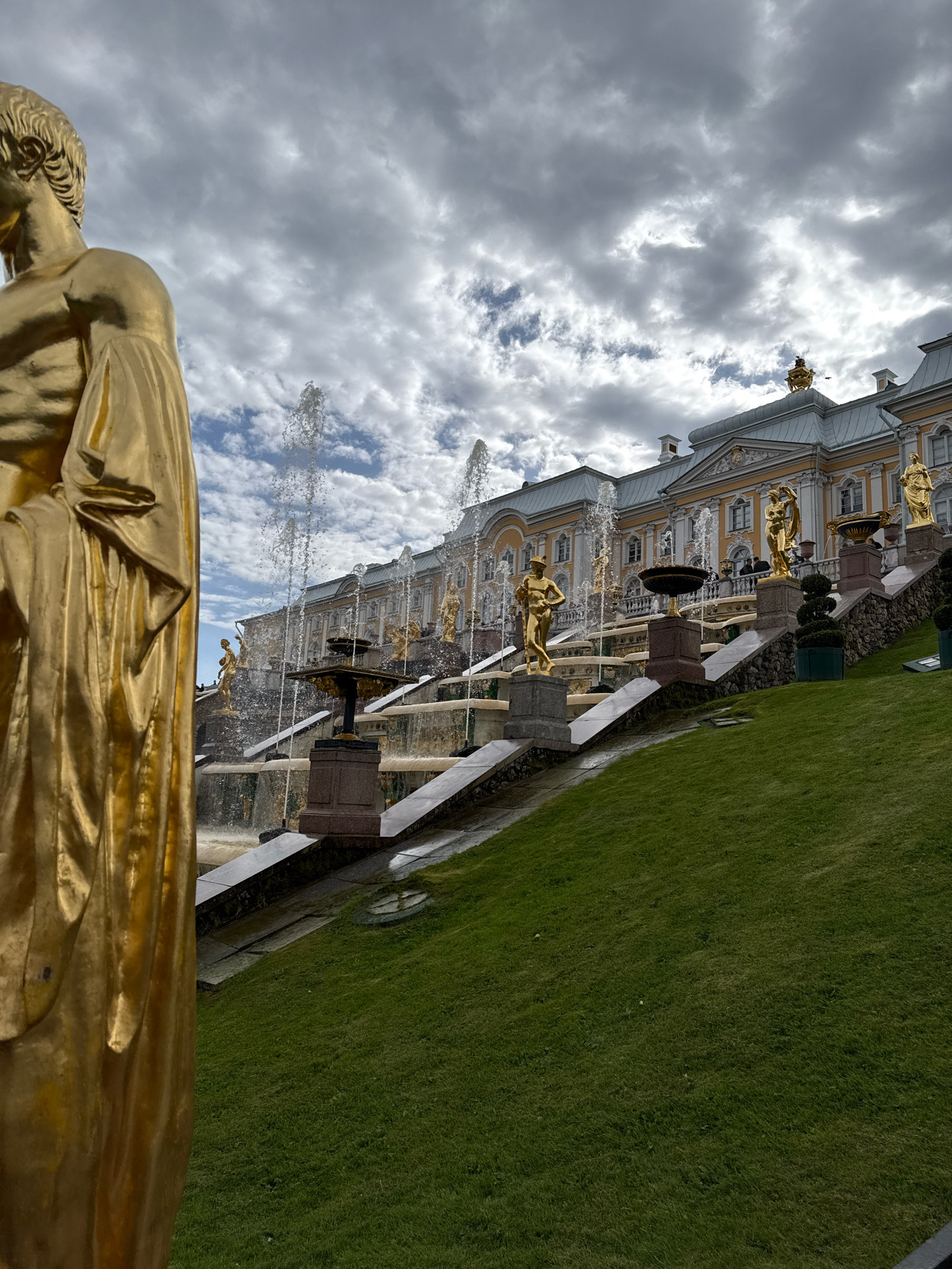 This image captures the Peterhof Grand Cascade in Peterhof, Russia, a magnificent fountain system leading to the Grand Palace. In the foreground on the left, a large, golden statue of a male figure, possibly a god or hero, is partially visible, its form rendered in a draped garment. Behind the statue, the Grand Cascade unfolds, a series of tiered water basins and sculptures adorned with golden figures. Water spouts upwards from numerous points within the cascade, creating a dynamic and shimmering effect. Several golden statues of mythological figures stand on pedestals amidst the flowing water. The cascade leads up to the yellow and cream-colored facade of the Grand Palace, which features intricate architectural details and is topped with a grey roof. The scene is set against a sky filled with dramatic, cloudy formations. A meticulously maintained green lawn slopes downwards from the cascade, adding to the grandeur of the setting.