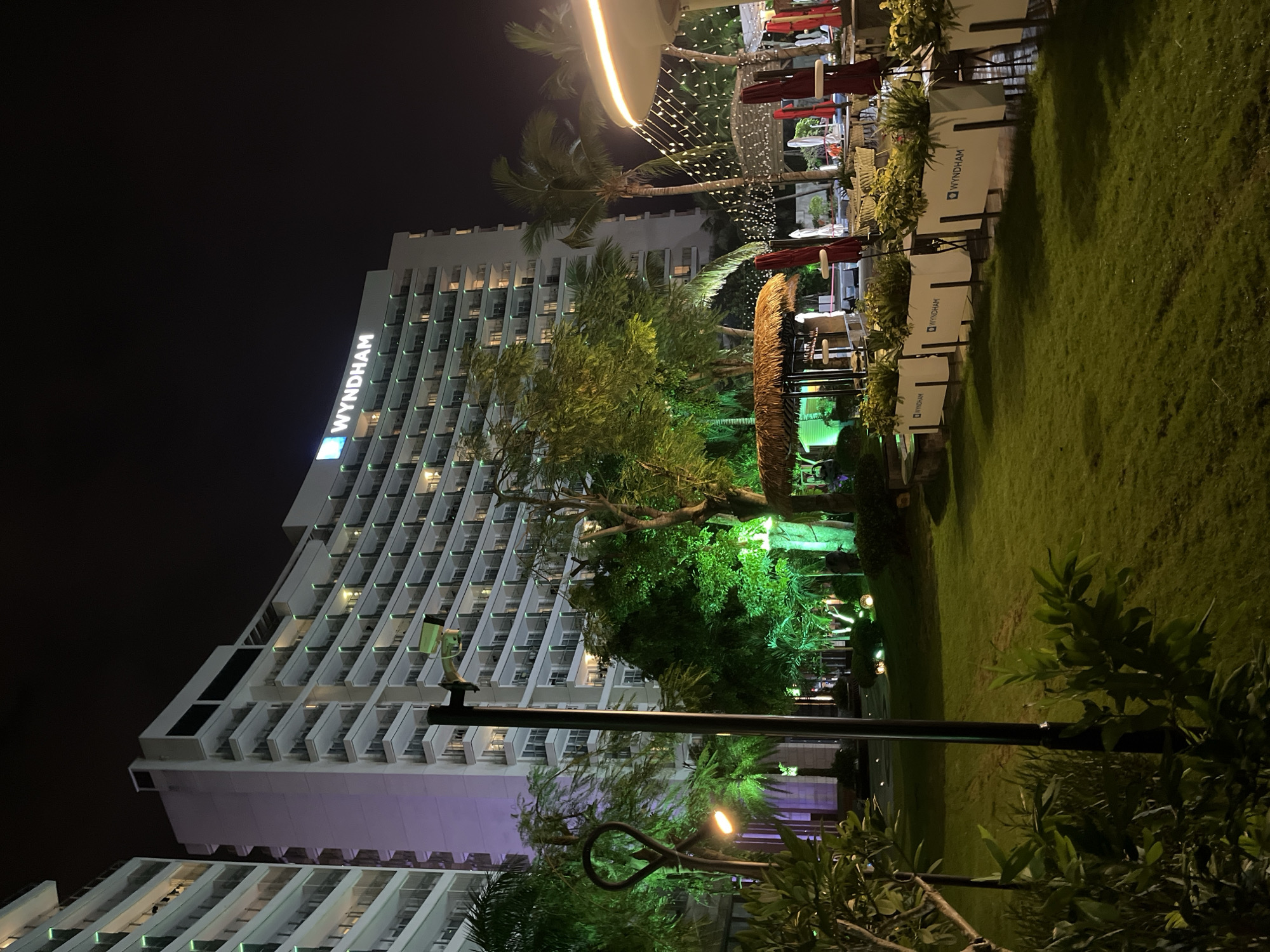 This media file depicts a nighttime scene at a resort, likely in Tianya District, China. The prominent subject is a large, modern hotel building with multiple floors and balconies, illuminated by interior lights. The "WYNDHAM" logo is clearly visible on the upper portion of the hotel.
In front of the hotel, an outdoor seating area with tables, chairs, and red umbrellas is set up on a well-maintained lawn. String lights are draped across the area, creating a festive ambiance. Palm trees and other lush greenery are scattered throughout the scene, some of which are illuminated with green lights. The overall setting suggests a resort or a luxury hotel with outdoor recreational facilities.
The time of day is clearly nighttime, indicated by the dark sky and artificial lighting. The weather appears clear, with no visible signs of rain or wind. The atmosphere is tranquil and inviting, suggesting a relaxing evening at the resort. There are no people visible in the image, focusing the attention on the architecture and the landscape. The scene is well-lit, highlighting the modern architecture of the hotel and the tropical vegetation. The presence of Wyndham branding suggests this is likely a hotel associated with that chain.