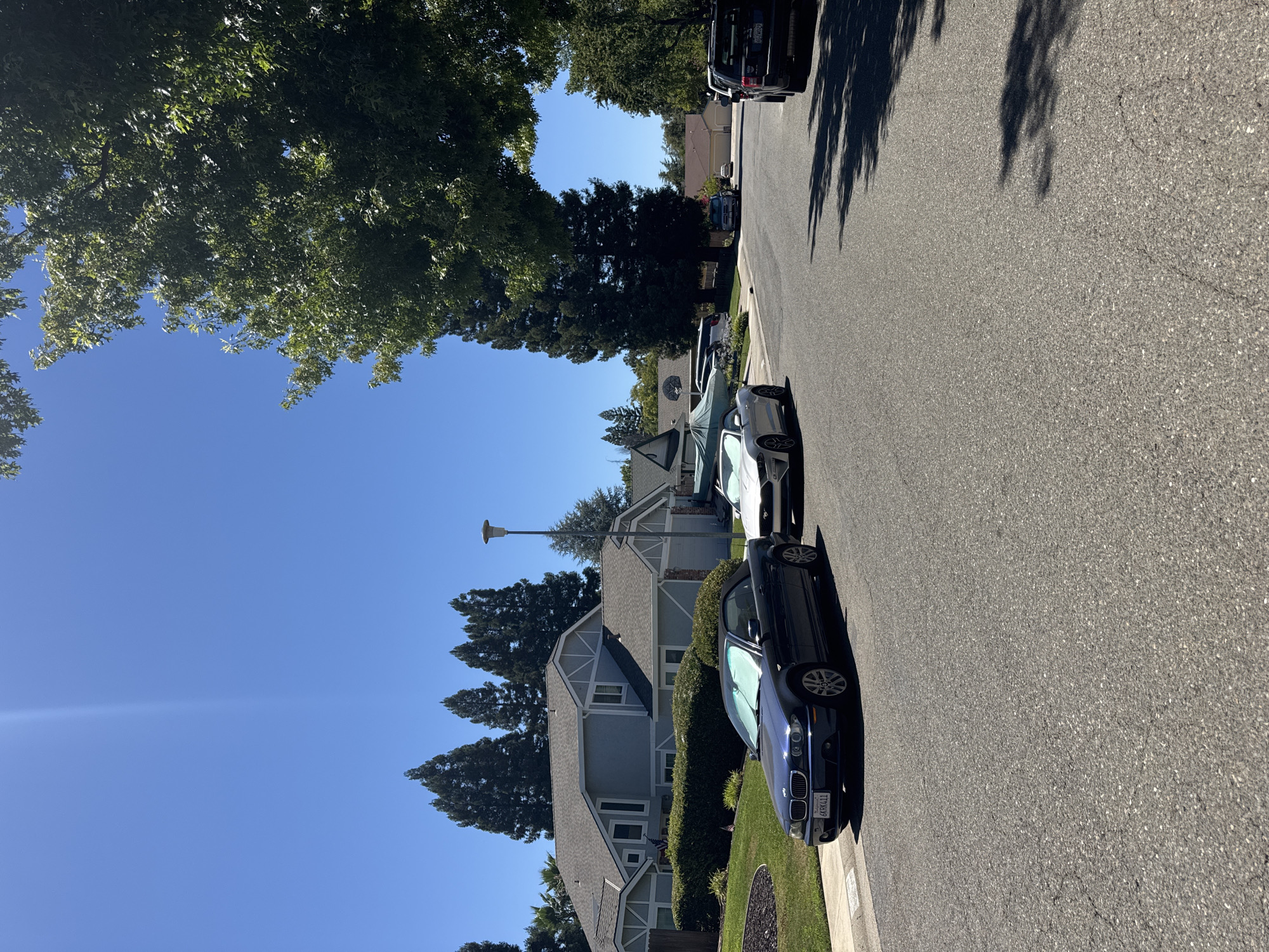 A suburban street scene in Citrus Heights, California, on a clear, sunny day. A dark blue convertible BMW 3 Series is parked on the left side of the asphalt road, facing away from the camera. A silver Ford Mustang convertible is parked to its right, also facing away. Behind the cars, a light blue house with a gray roof and white trim is visible, surrounded by green bushes and a manicured lawn. Tall, dark green cypress trees line the background, with a large, leafy green tree canopy arching over the right side of the road from the foreground. A lamppost stands to the right of the blue house. Further down the street, more houses and trees are visible. Shadows from the trees are cast across the road. A dark-colored SUV is parked further down the street on the right.