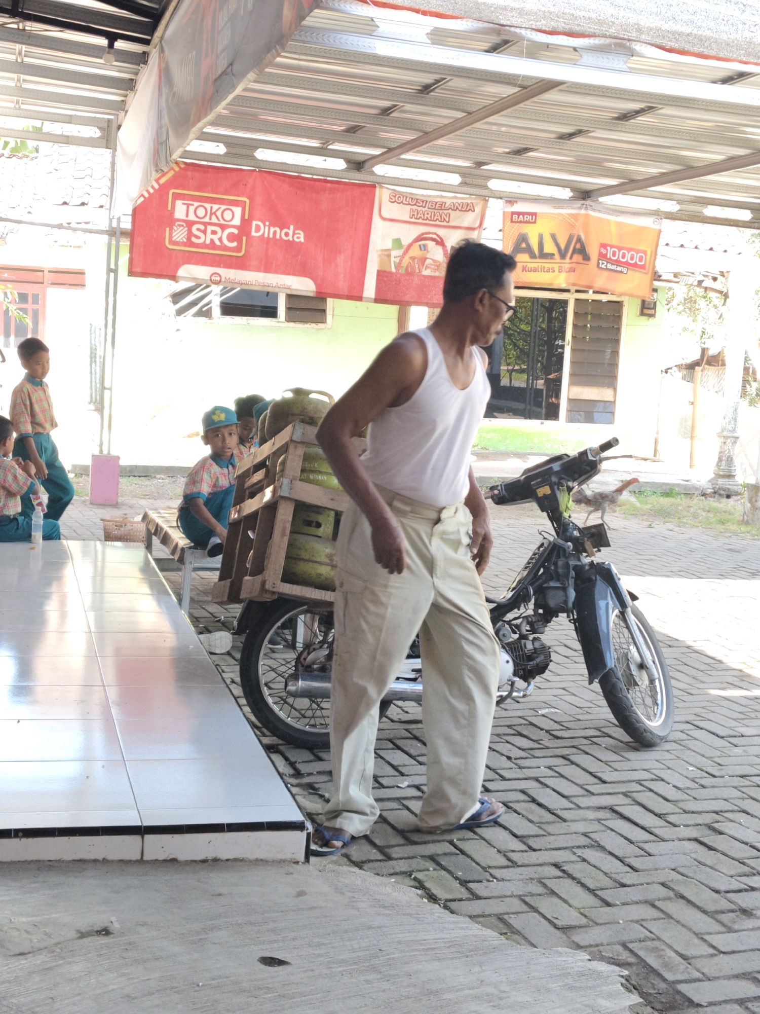 In Bangsalsari, Indonesia, a man wearing a white tank top and tan pants stands beside a motorcycle laden with gas canisters. The motorcycle is parked on a brick-patterned pavement under a covered structure with a corrugated metal roof. To the left, three young boys in school uniforms are seated on a bench near a tiled floor. One boy, wearing a blue cap, is looking towards the camera. Behind the boys, a pink object, possibly a stool, is visible. In the background, there are shop signs, one red with "TOKO SRC Dinda" and another orange with "ALVA Rp 10.000". A chicken is seen walking on the ground near the front of the motorcycle. The scene appears to be outdoors, likely in front of a small shop or market.