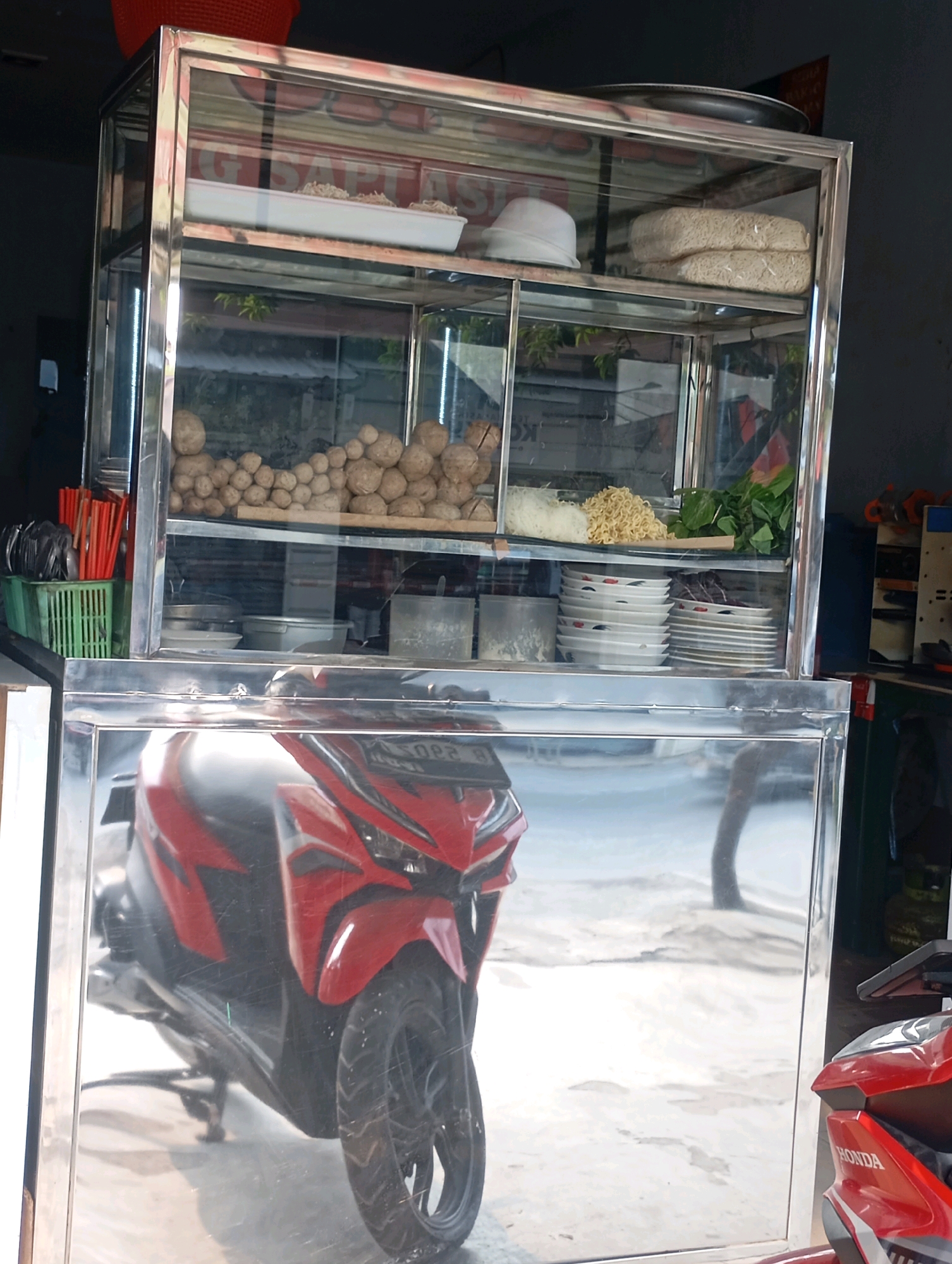 A street food stall is depicted, featuring a two-tiered glass and stainless steel display case filled with food items and serving dishes. The upper shelf contains what appears to be meatballs, noodles, and leafy greens. The lower shelf holds stacks of plates. A red motorcycle is prominently reflected in the mirrored base of the stall. The stall is situated outdoors, with a concrete ground visible, suggesting a location within a city. A red basket is partially visible at the top left of the image. The overall scene suggests a typical food vendor setup in a public area. The location is identified as Babelan, Indonesia.