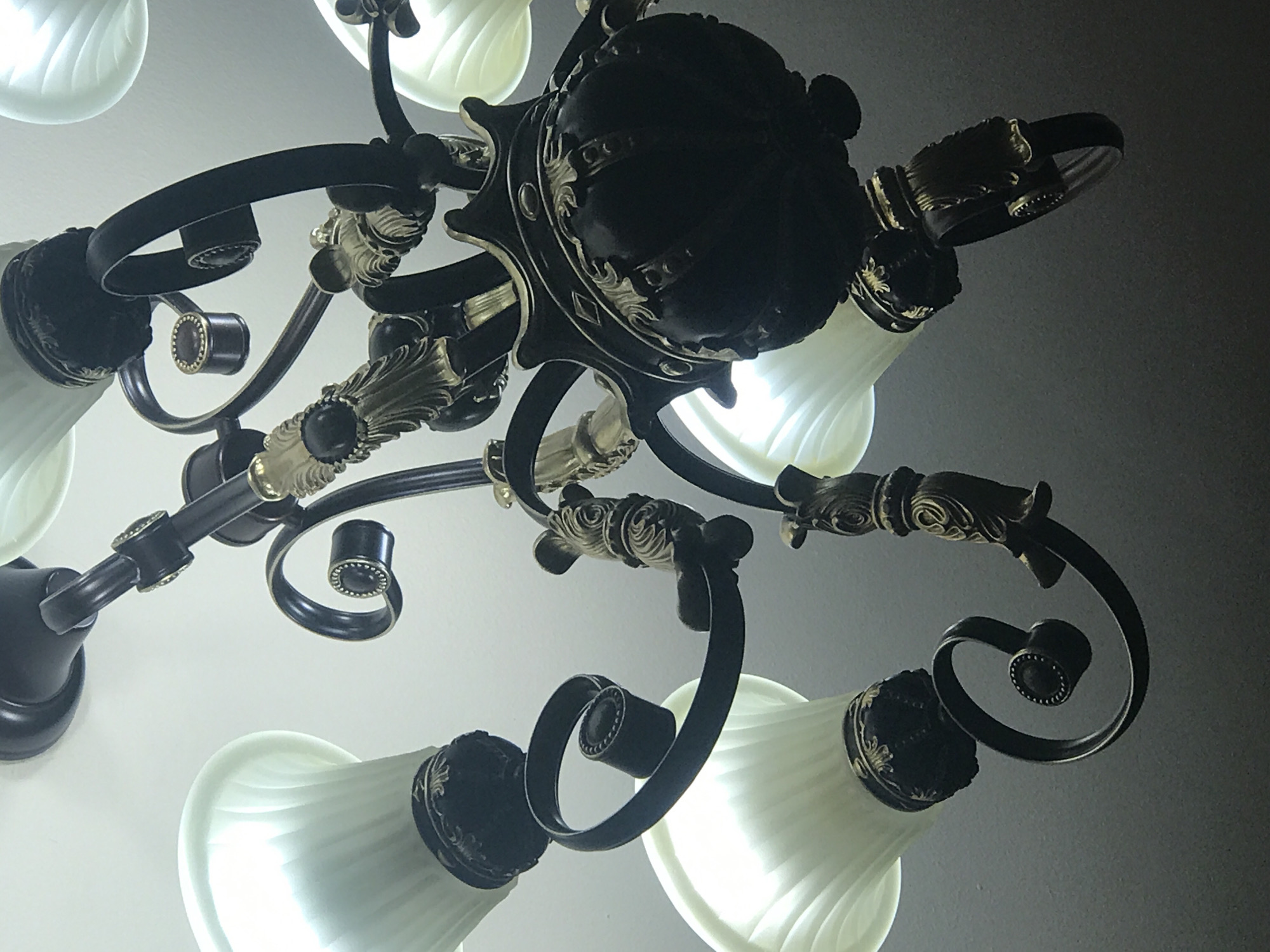 A low-angle shot reveals a chandelier with black and gold accents and multiple frosted glass shades. The chandelier's central structure is dark and ornate. The light shades are a soft white, and the bulbs within are illuminated. The background transitions from a bright white near the lower left to a gradient of gray towards the upper right. The photo was not taken in Yerevan, Armenia.