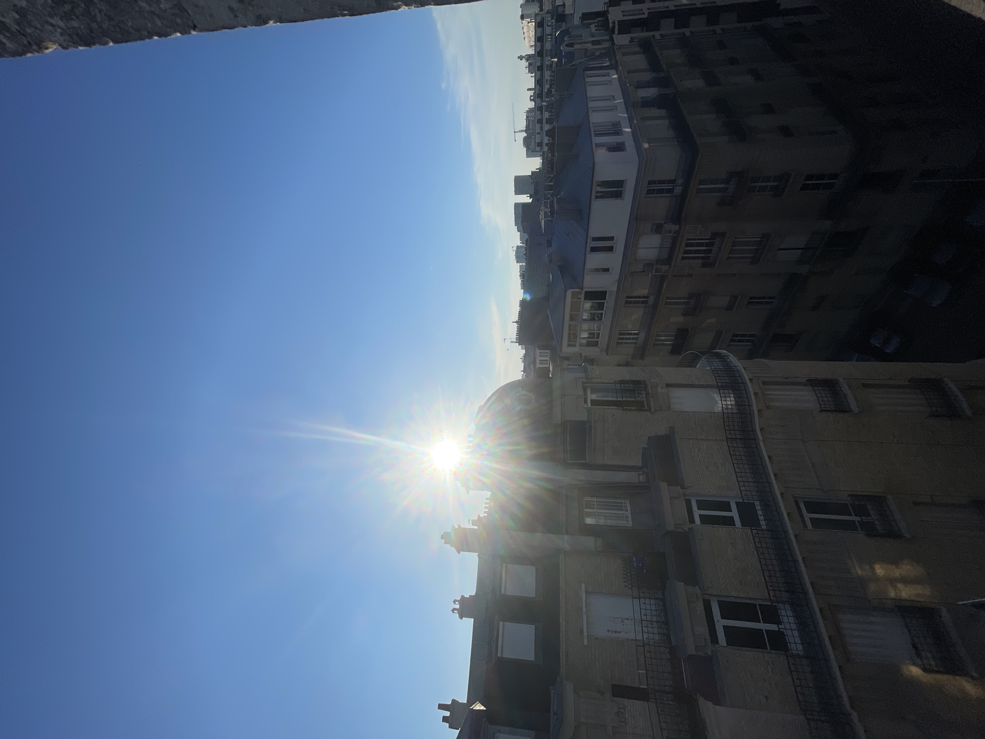The image captures a bright, sunny day over the rooftops of Paris, France. The sun is prominently featured, creating a starburst effect with its rays, positioned in the upper center of the frame against a clear blue sky. In the foreground, on the left, is the side of a building with tan brickwork, featuring multiple windows with white frames and balconies with black railings. A rounded, domed structure, possibly part of a building or monument, is visible on this building, partially obscured by the sun's glare.
To the right and slightly further back, another building with a light-colored facade and blue-grey roofs is visible. This building displays a series of windows and balconies. Beyond these foreground buildings, a cityscape of Parisian architecture unfolds, with a mix of taller and shorter buildings extending towards the horizon. Cranes are visible in the distance, suggesting ongoing construction in the city. The perspective suggests the photo was taken from a high vantage point, possibly a window or balcony of a building. The overall impression is one of an urban panorama under a clear and radiant sky.