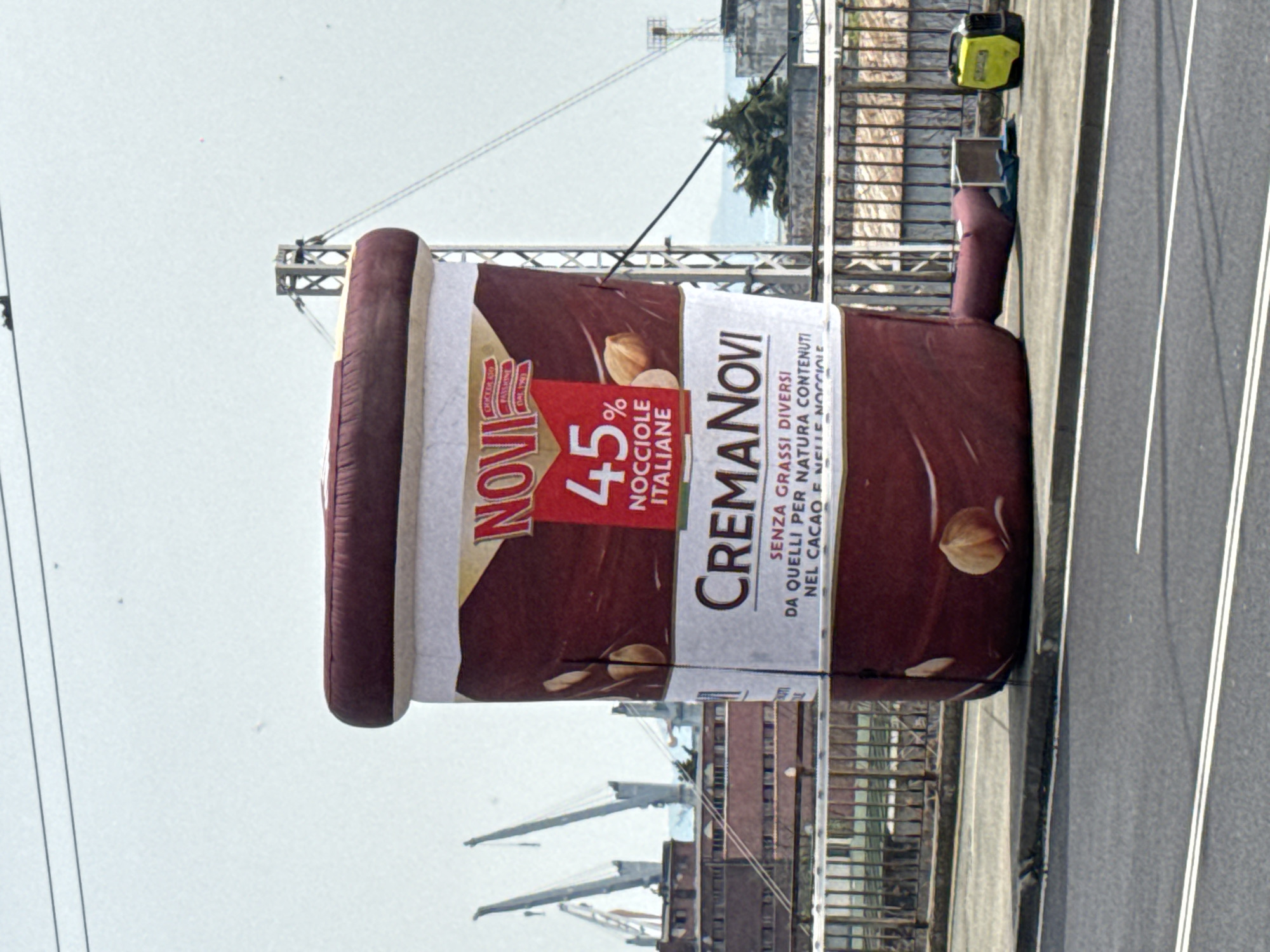 The image shows a large inflatable advertisement for "Crema Novi," a hazelnut spread, standing on a paved surface. The advertisement, shaped like a jar, displays the Novi brand logo, the text "45% NOCCIOLE ITALIANE," and descriptive text about the product's ingredients. The environment includes a road with white lane markings, a metal railing, and buildings in the background. Cranes are visible in the distance, implying an industrial or port area. A yellow generator and equipment case are on the ground near the inflatable. Based on the presence of cargo cranes in the background, it is likely located near the Port of Burgas in Bulgaria.