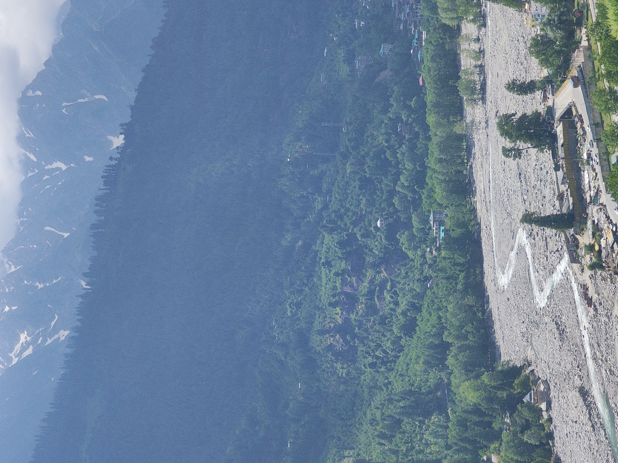 The image depicts a wide view of a mountainous landscape with a river flowing through a valley. In the foreground, a dry riverbed filled with large grey stones is visible, with a narrow, winding river of turquoise water flowing through it. Several structures are clustered near the river, including small buildings, a bridge, and what appears to be a construction site with scaffolding and orange trucks. The riverbanks are lined with lush green trees, and further up the valley, the slopes are covered in dense forests. In the distance, smaller buildings and houses are scattered across the hillsides, suggesting a populated area within the valley. The background is dominated by a towering mountain range, with its peaks partially obscured by clouds and snow. The overall scene conveys a sense of natural beauty and a remote, mountainous settlement, likely in the region of Vashisht, India, known for its similar geographical features and proximity to the Himalayas. There are no visible people in the image.