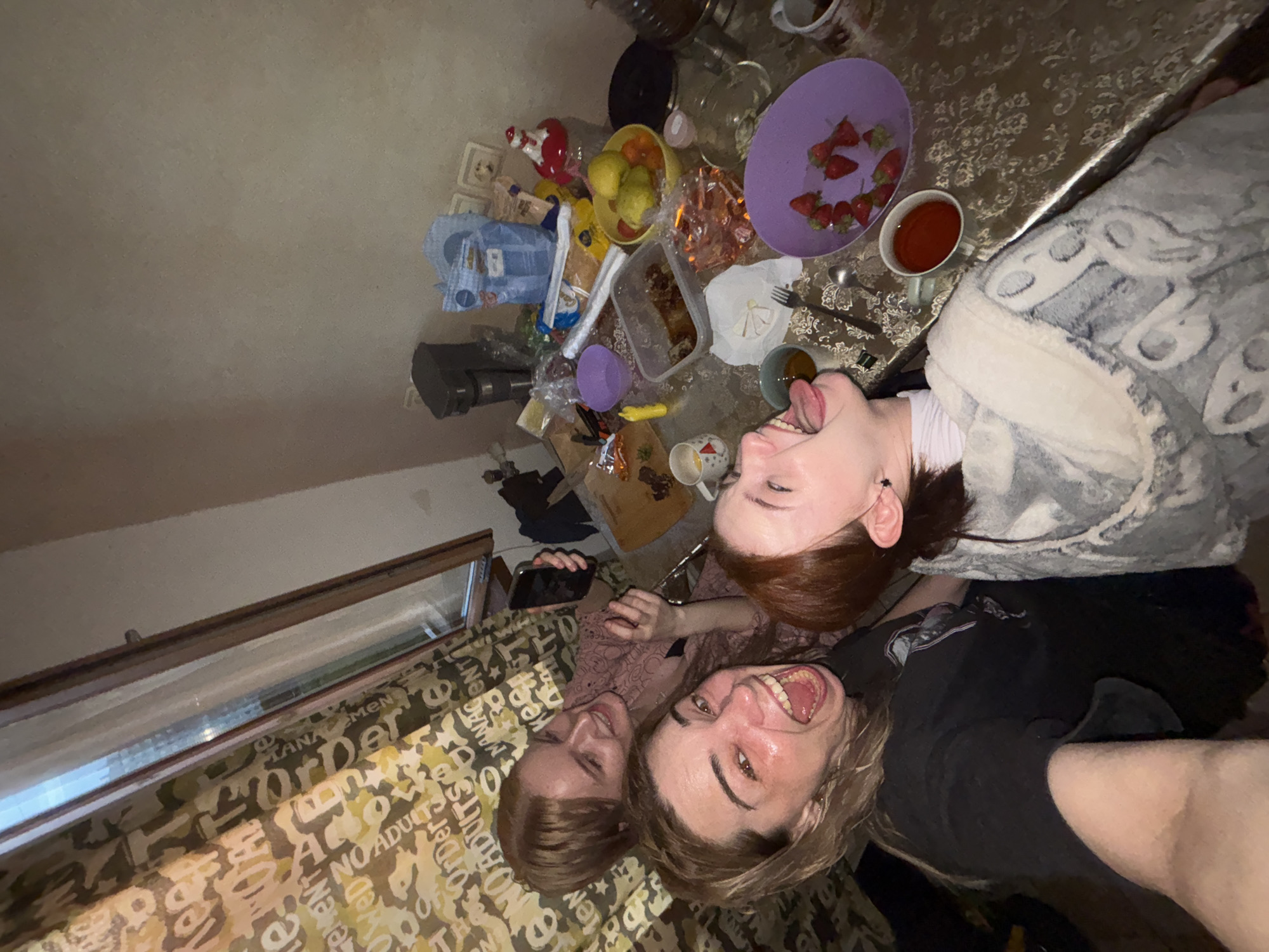 The image captures three young women in an indoor setting, likely a casual home environment, engaged in a playful, relaxed moment. The photograph is taken from a high angle, looking down upon the subjects, who appear to be lying down or leaning back.
Closest to the camera, a woman with light brown hair smiles broadly with her mouth open, her face appearing slightly flushed. Beside her, another woman with reddish-brown hair playfully sticks out her tongue and squints her eyes, conveying a sense of fun. She is wearing a light-colored top and is partially covered by a grey blanket with a patterned design. Further back and to the left, a third woman with brown hair holds a smartphone and looks towards the camera with a gentle smile.
The women are positioned around a table covered with a patterned tablecloth featuring brown and lighter designs. The table is laden with various items, suggesting a casual meal or snack time. Visible objects on the table include a purple plate filled with sliced strawberries, a mug containing a dark red beverage (possibly tea or juice), a yellow bowl with fruit (appearing to be lemons or limes), and a clear plastic container holding food. Other miscellaneous dishes, bottles, and bags contribute to a somewhat cluttered, lived-in atmosphere. In the background, a plain, light-colored wall is visible, along with a window that has patterned curtains beneath it. The curtains feature repetitive text, with discernible words such as "Order," "MEN," "NO ADULT," and "LOVE."
The indoor lighting suggests the time of day is likely evening or night. The overall emotion conveyed by the subjects is one of joy, relaxation, and camaraderie.