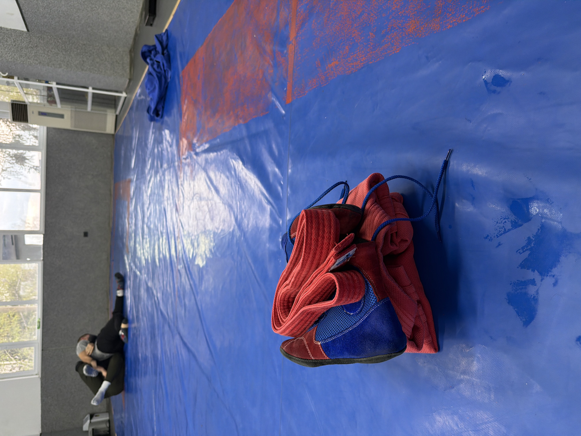 The image captures an indoor sports facility, likely a wrestling or martial arts gym, in Panagyurishte, Bulgaria. The floor is covered with a large, royal blue mat, which shows signs of use and has faded red rectangular markings, possibly indicating boundaries or specific areas for practice.
In the foreground, a pair of red and blue wrestling shoes with blue laces rests on the mat. Next to the shoes is a neatly folded red piece of athletic clothing, possibly a wrestling singlet or part of a uniform. Further back on the mat, a crumpled blue piece of clothing is visible near a wall.
On the far left side of the frame, a person is on the mat, facing away from the camera. They are wearing dark pants, a dark long-sleeved top, a grey cap or head covering, and white socks. The person is in a low, crouched or kneeling position, suggesting they might be stretching, resting, or preparing for an activity.
The setting includes a large window on the left wall, letting in natural light and showing glimpses of trees outside. Below the window, an air conditioning unit is mounted. The walls are a neutral light grey. The overall impression is that of a functional training space, likely captured during a quiet moment before or after a practice session. No discernible text is visible in the image.